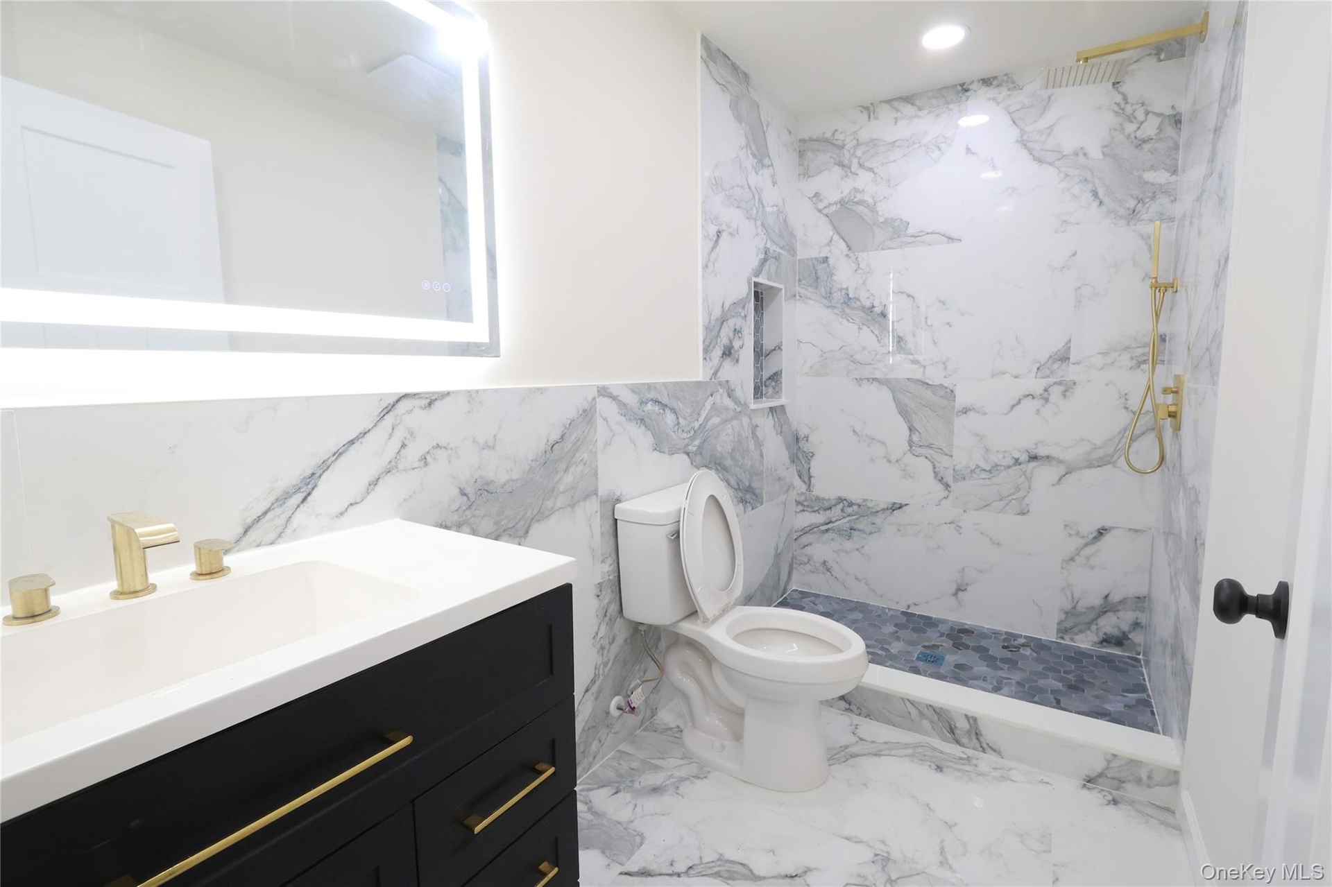 Vanity with integrated sink and gold-tone fixtures, backlit mirror, floor-to-ceiling marbled tile, and a walk-in shower with a recessed shelf
