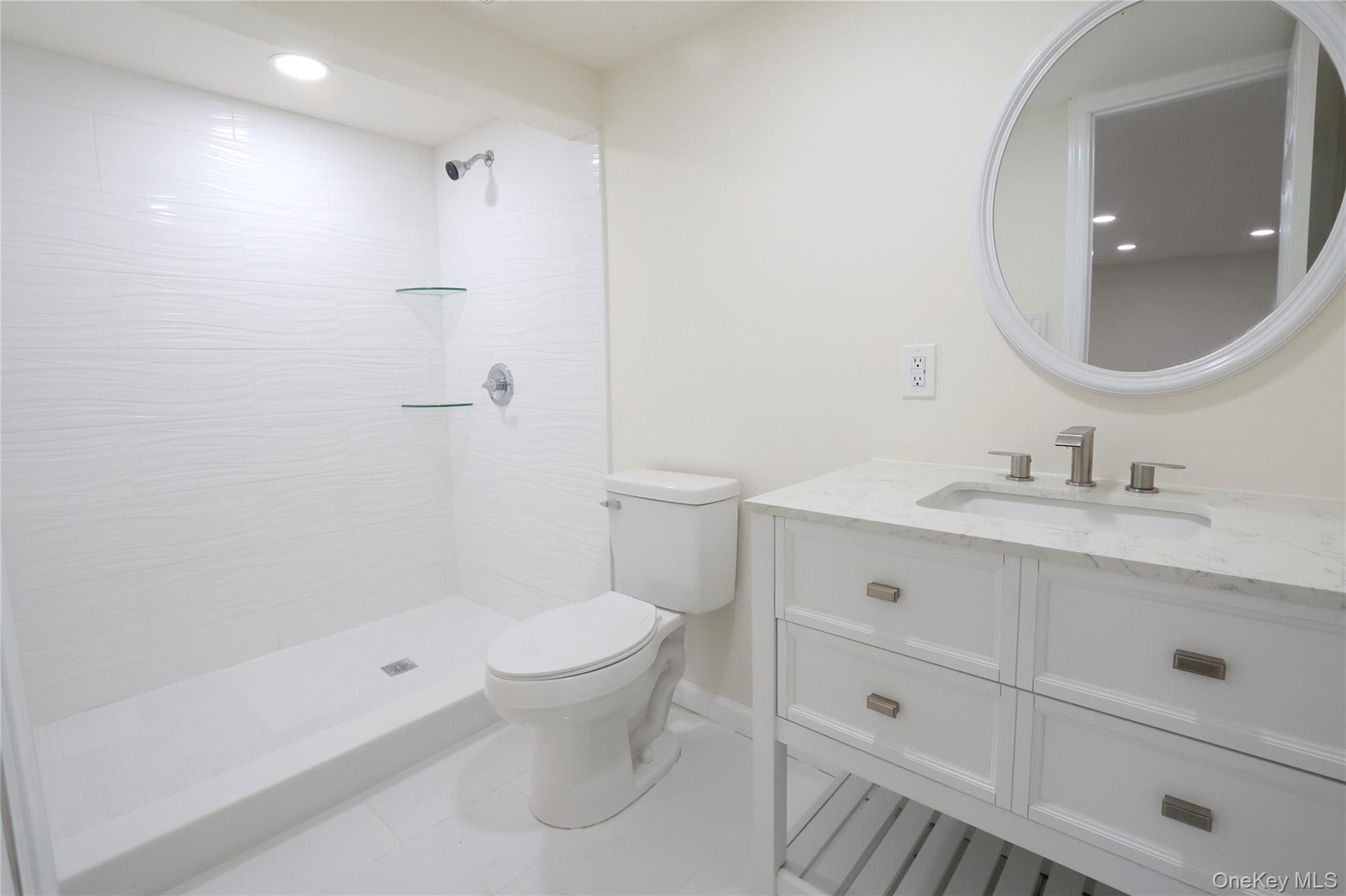 Bathroom featuring a white vanity with a stone countertop, a circular mirror, a walk-in shower with textured wall tiles, and glass corner shelves