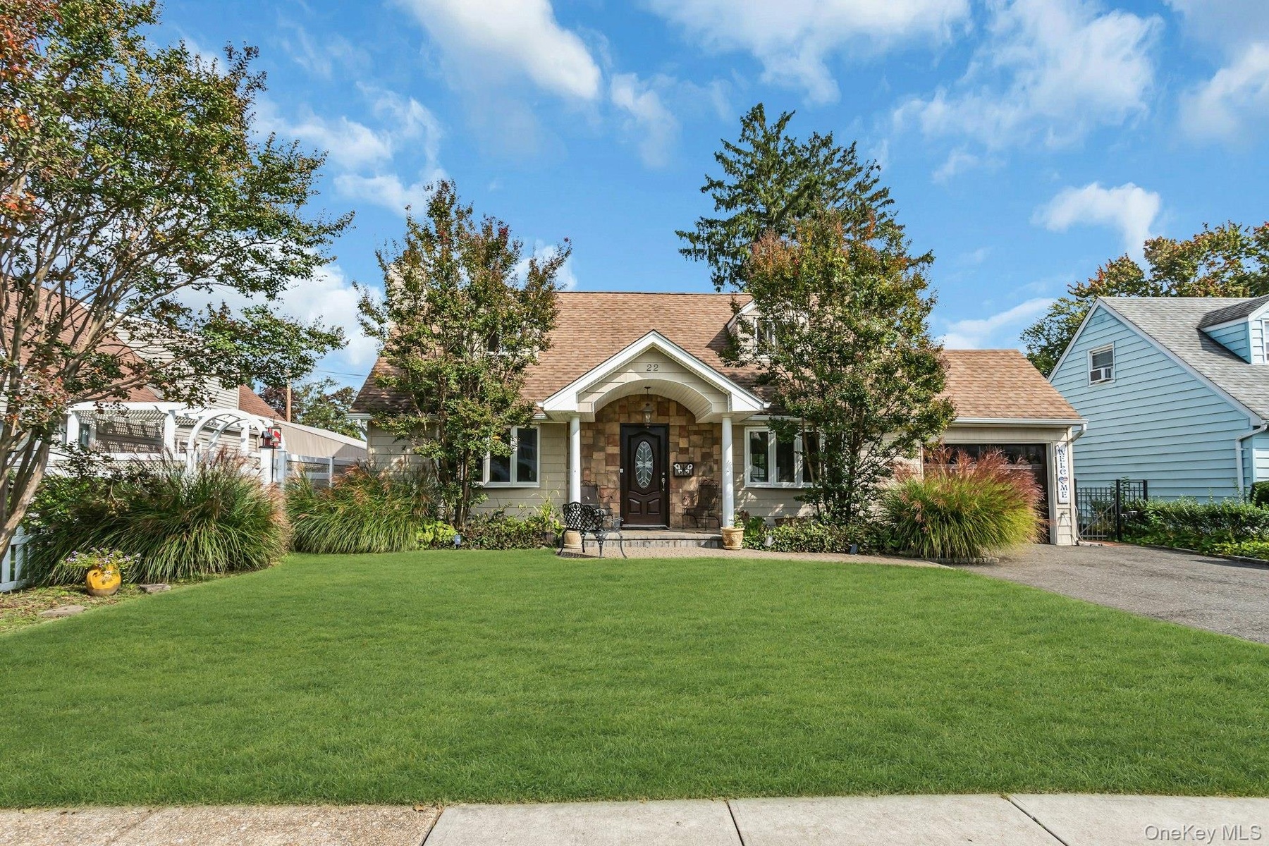 Pitched roof with asphalt shingles, stone facade entrance, attached garage with wood-finish door, paved driveway, and established landscaping