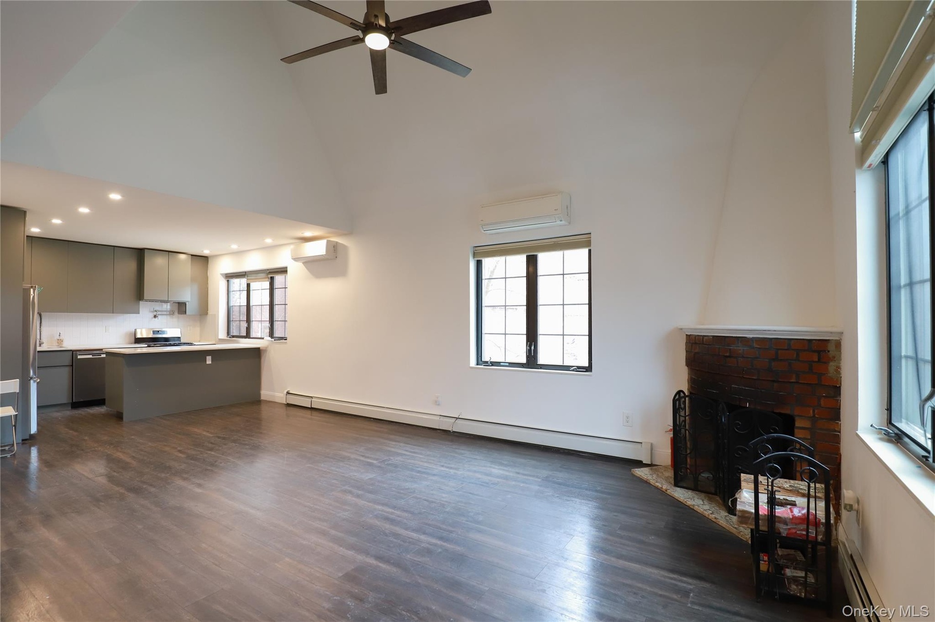 Vaulted ceiling with a ceiling fan, wood-finish flooring, a brick fireplace, modern kitchen cabinetry, and stainless steel kitchen appliances
