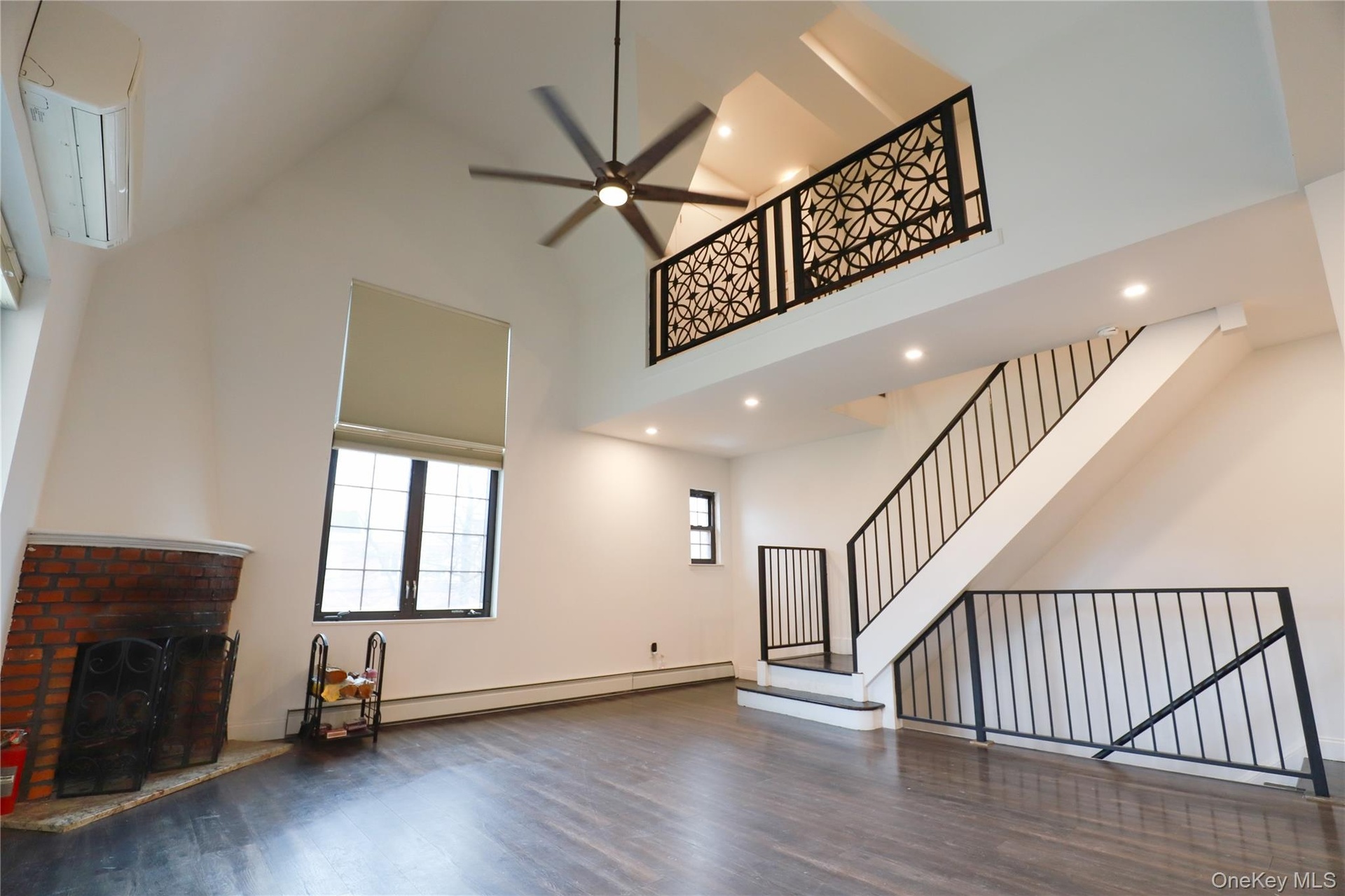 Vaulted ceiling with an exposed beam, brick fireplace with a stone hearth, dark wood-finish flooring, and a loft with decorative ironwork railing