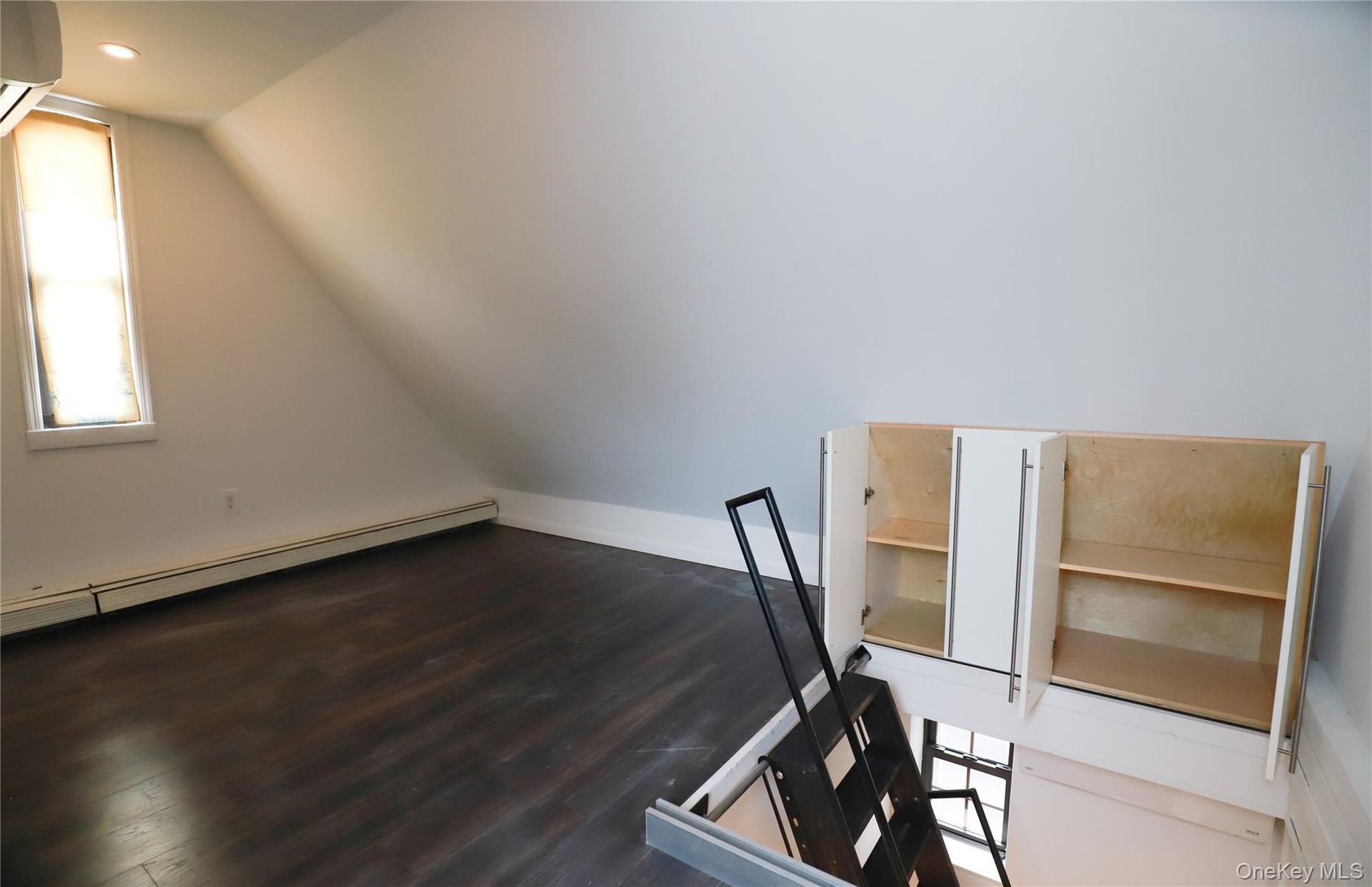 Dark wood-finish flooring with white baseboards, a single window with blinds, recessed ceiling lighting, built-in white cabinetry, and a black metal l