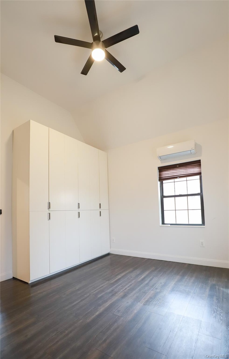 Vaulted ceiling with a ceiling fan, dark wood-finish flooring, built-in white cabinetry, window with blind, and a wall-mounted air conditioning unit