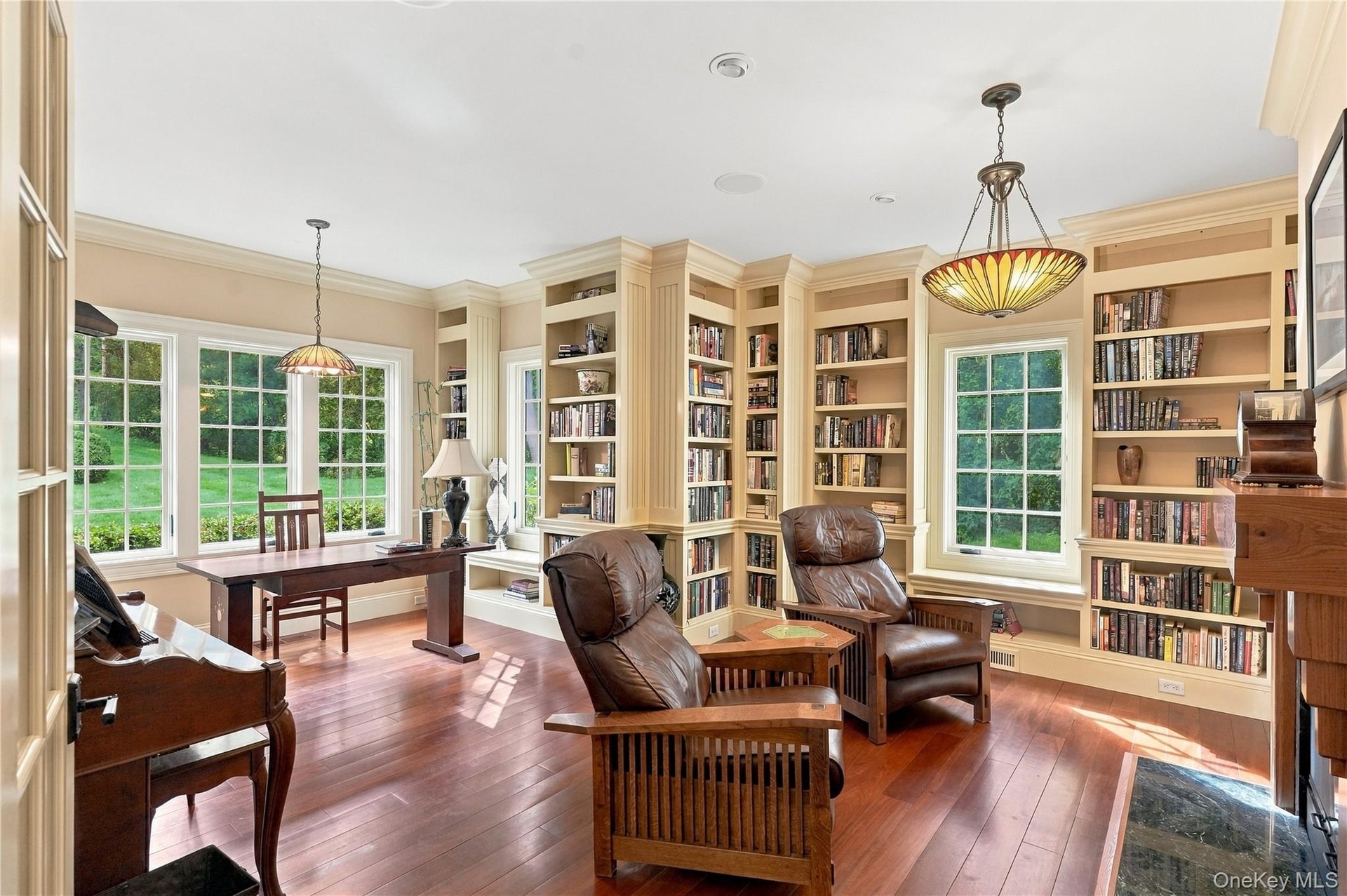 An elegant, sun-filled library featuring floor-to-ceiling custom built-in bookshelves, exquisite millwork, and expansive windows overlooking the lush 
