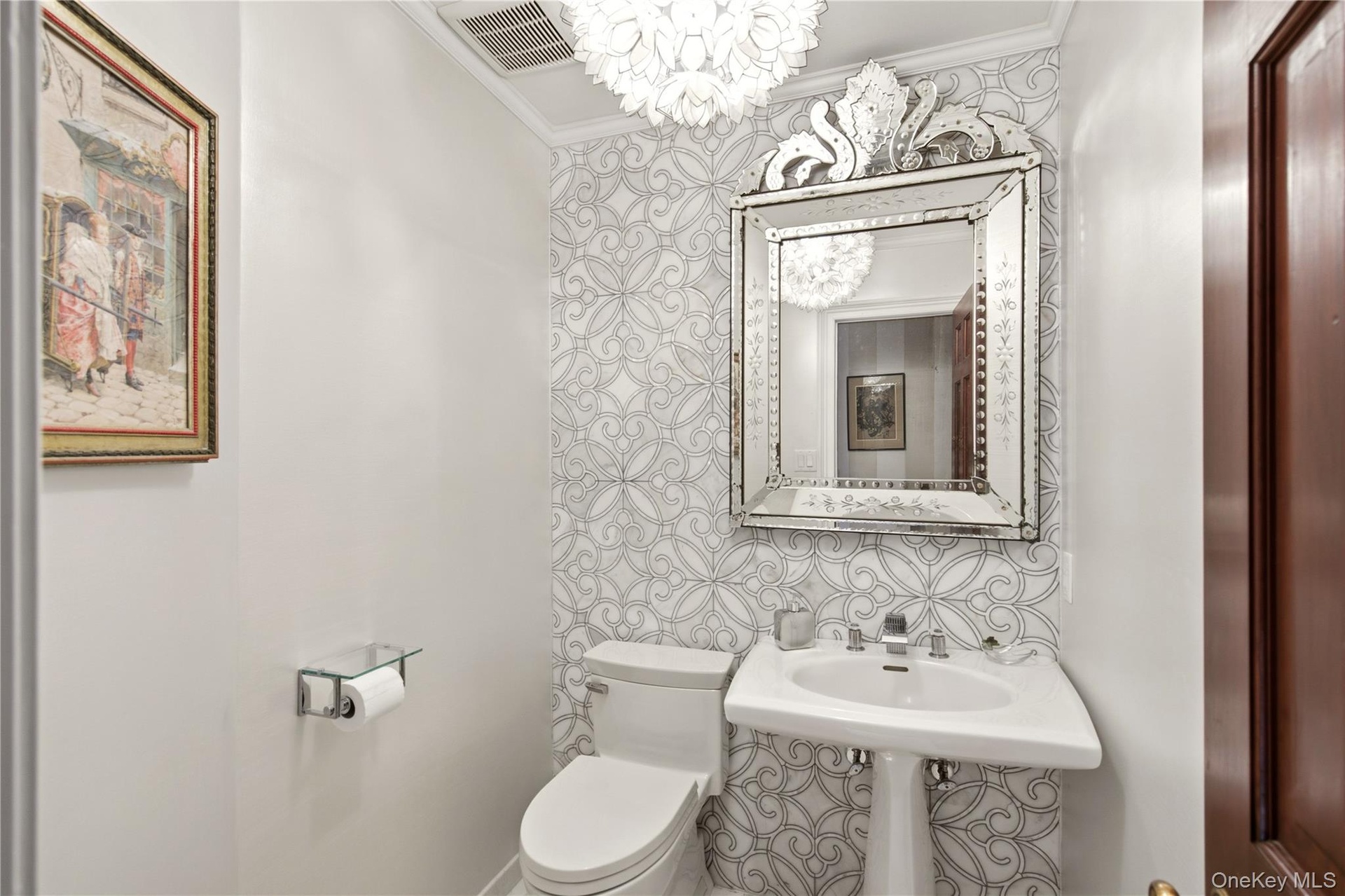 Pedestal sink with chrome fixtures, decorative wall-mounted mirror, patterned accent wall, and ceiling-mounted light fixture