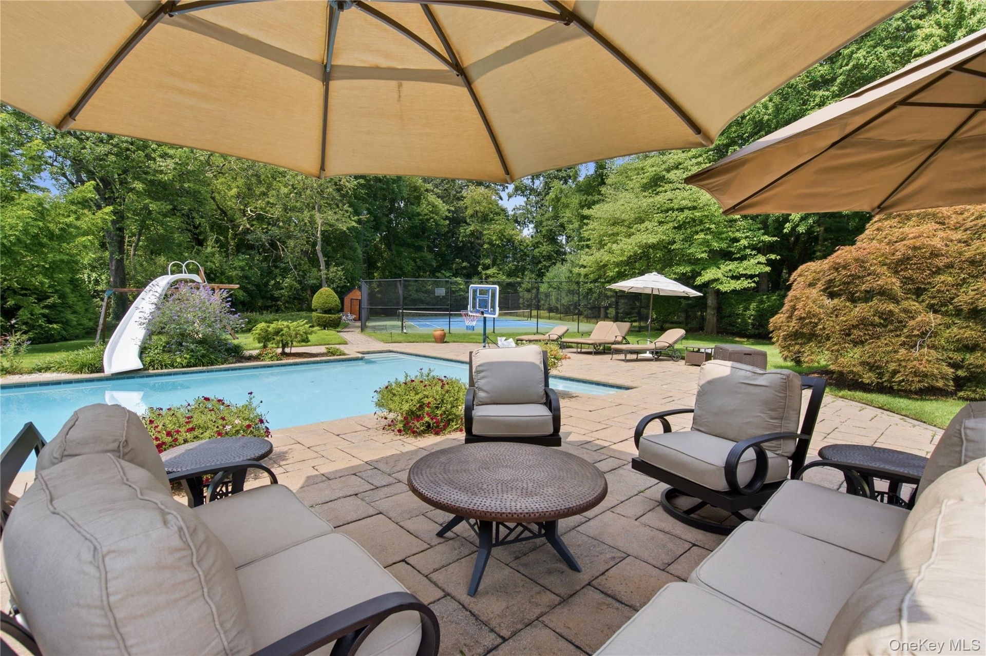 Paver patio with multiple shade umbrellas, a swimming pool with an attached slide, and a tennis court