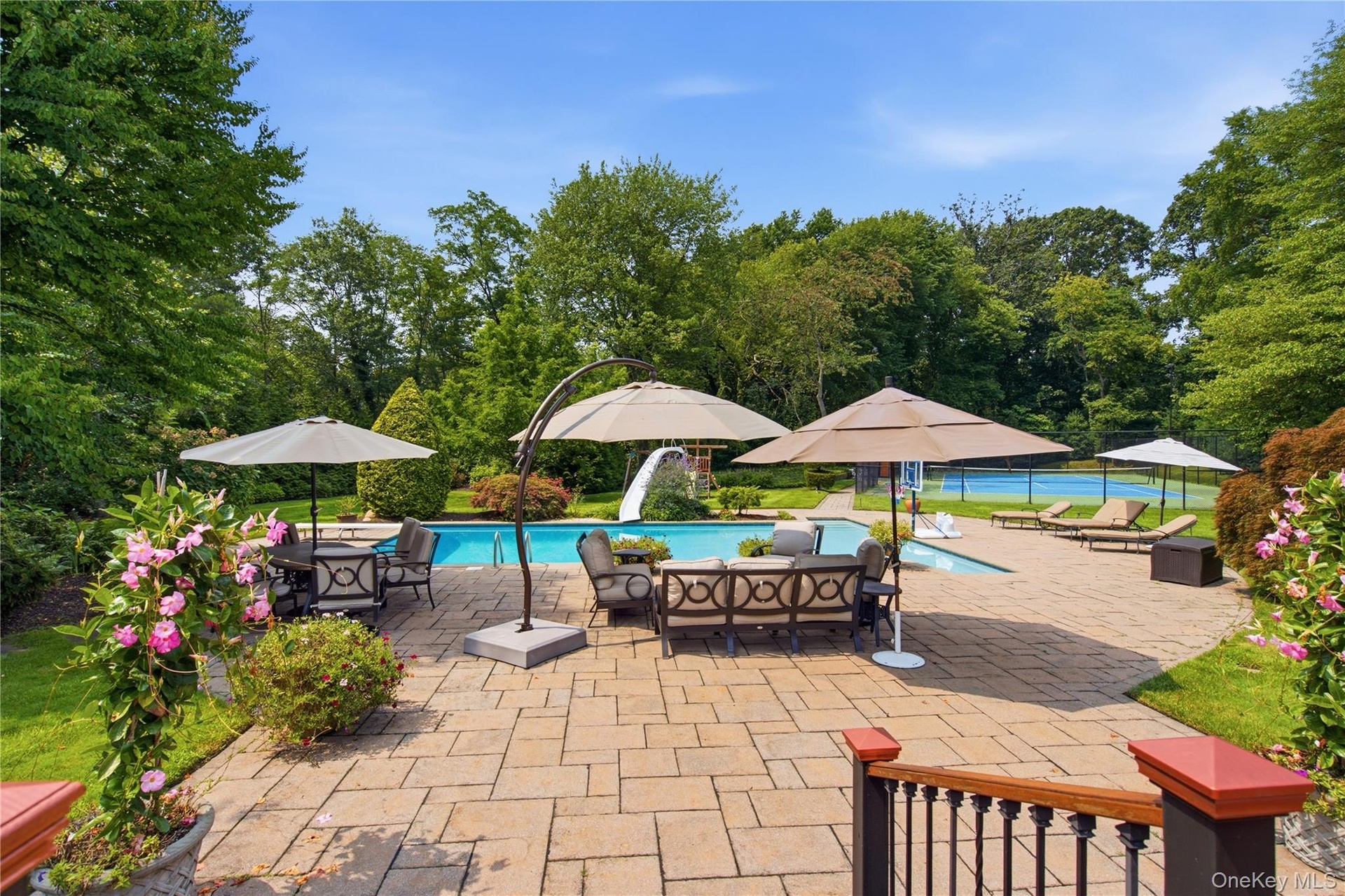 Expansive paver patio with an in-ground swimming pool, mature perimeter landscaping, and a fenced tennis court