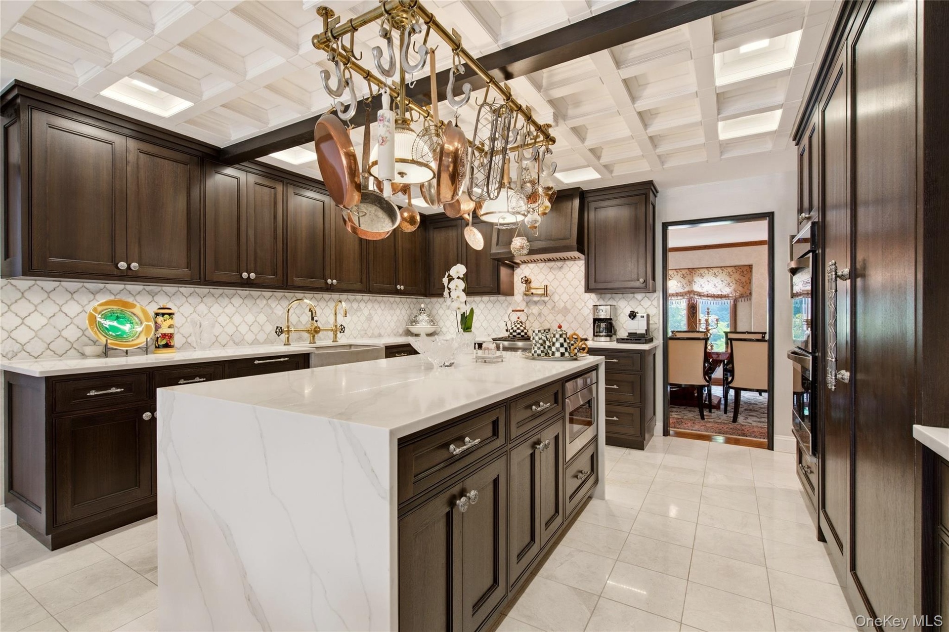 Black  oak wood cabinetry, a large center island with a waterfall countertop, tiled backsplash, coffered ceiling, and integrated stainless steel doubl