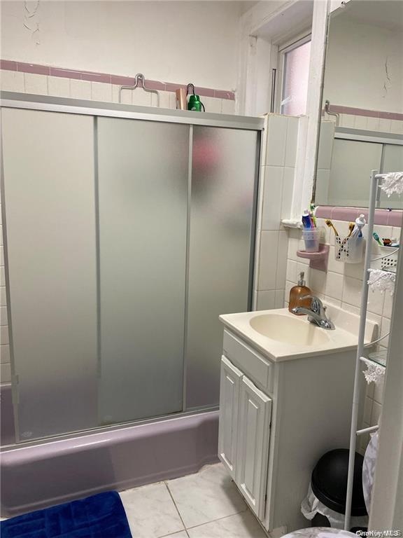 Tile flooring with white grout, a white vanity with a built-in sink and chrome faucet, and a shower/tub combination with sliding frosted glass doors