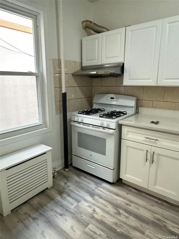 White shaker-style cabinetry, stainless steel range hood, tile backsplash, wood-finish flooring, and a white gas range
