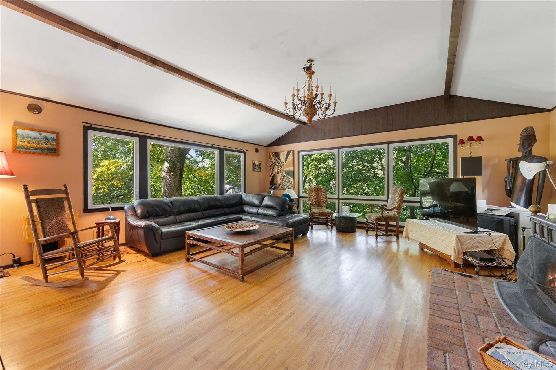 Vaulted ceiling with exposed wood beams, hardwood flooring, expansive window wall, brick fireplace, and a chandelier light fixture.