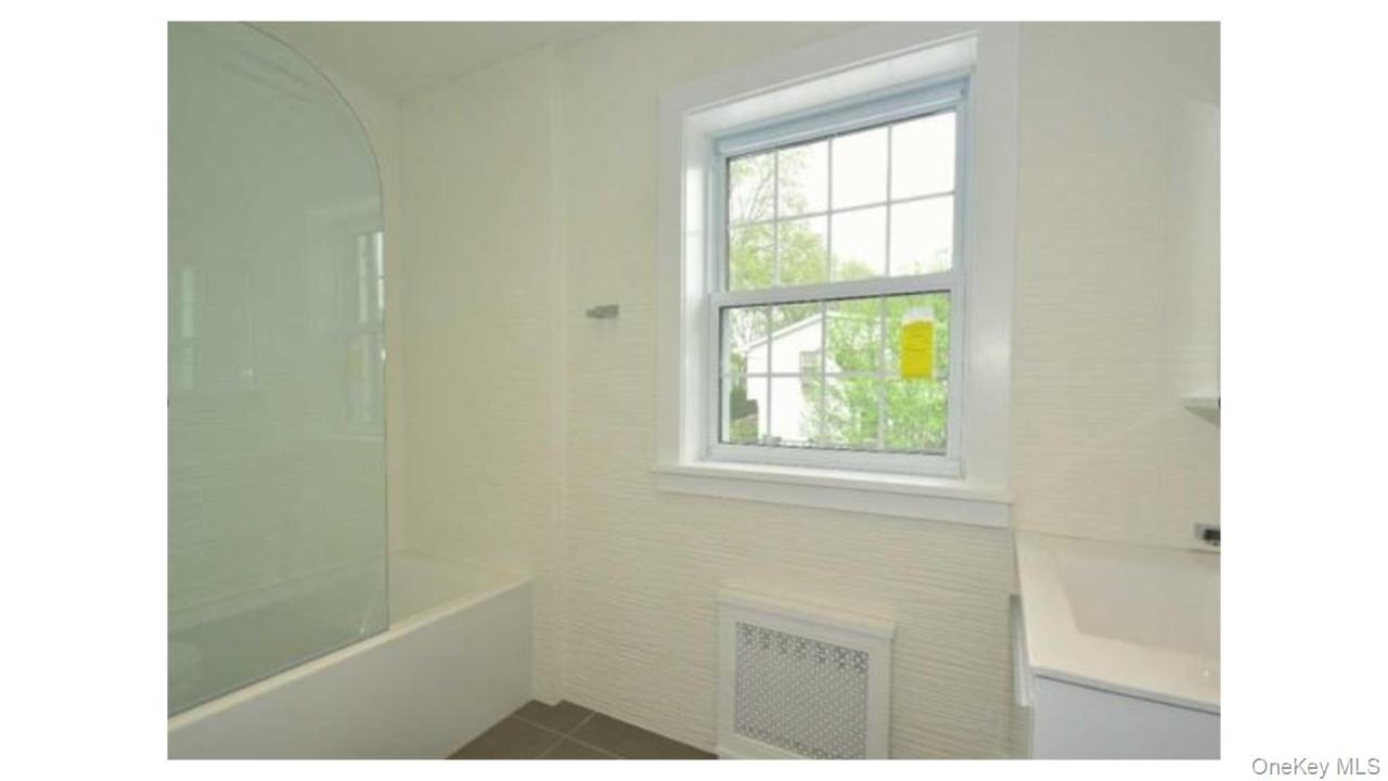 White bathtub with a curved glass shower partition, white window with grid panes, textured wall covering, and a white vanity with an integrated sink