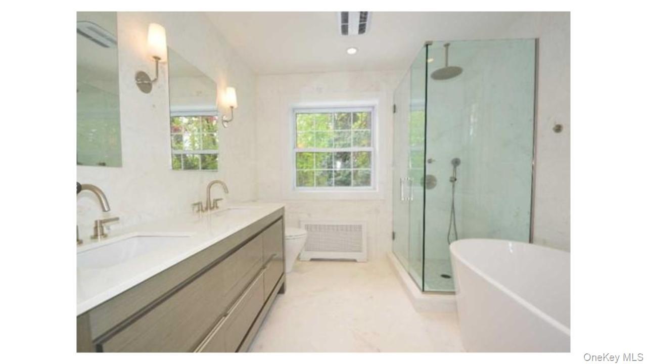 Dual vanity with integrated sinks, frameless glass shower enclosure, freestanding soaking tub, and natural light from a double-hung window