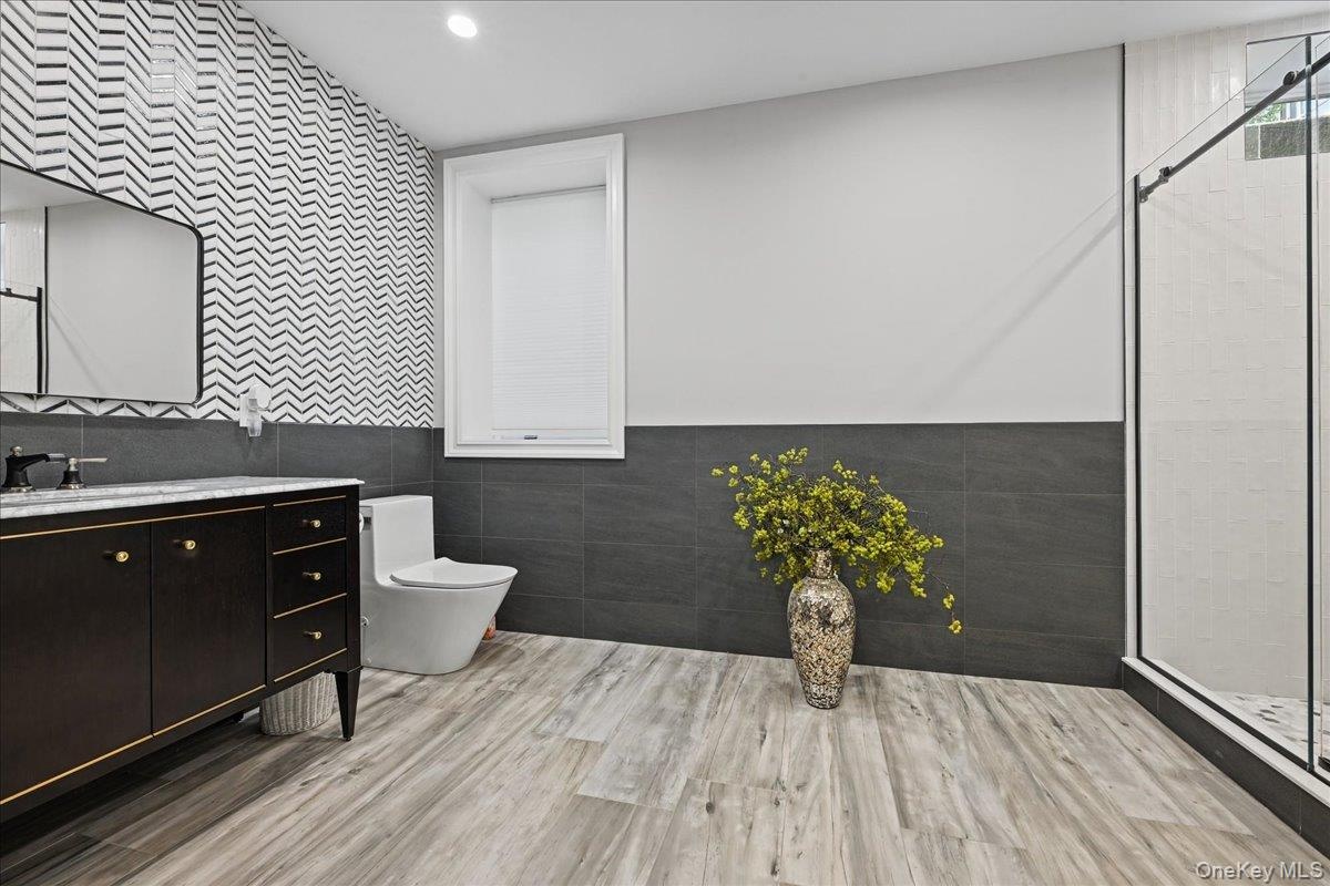 Modern bathroom featuring wood-finish flooring, a freestanding vanity with gold-tone hardware, a frameless shower enclosure, and a window with blinds