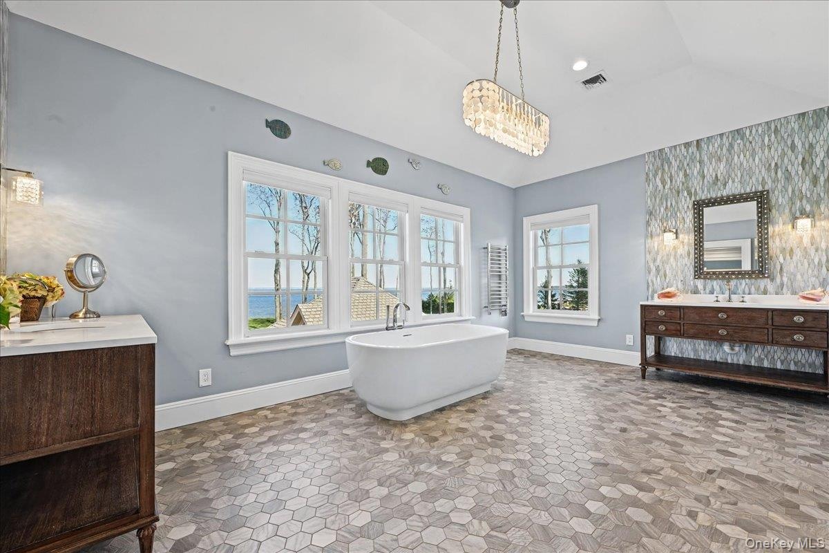Hexagonal tile flooring, freestanding soaking tub, dual wood-finish vanity with countertop, textural accent wall, and multiple windows