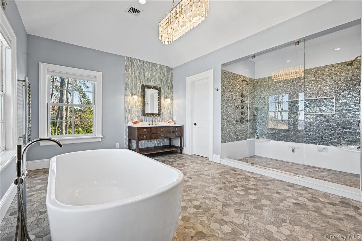 Freestanding soaking tub, walk-in shower with glass enclosure, wood-finish vanity with dual sinks, mosaic tile accent wall, and hexagonal floor tiling