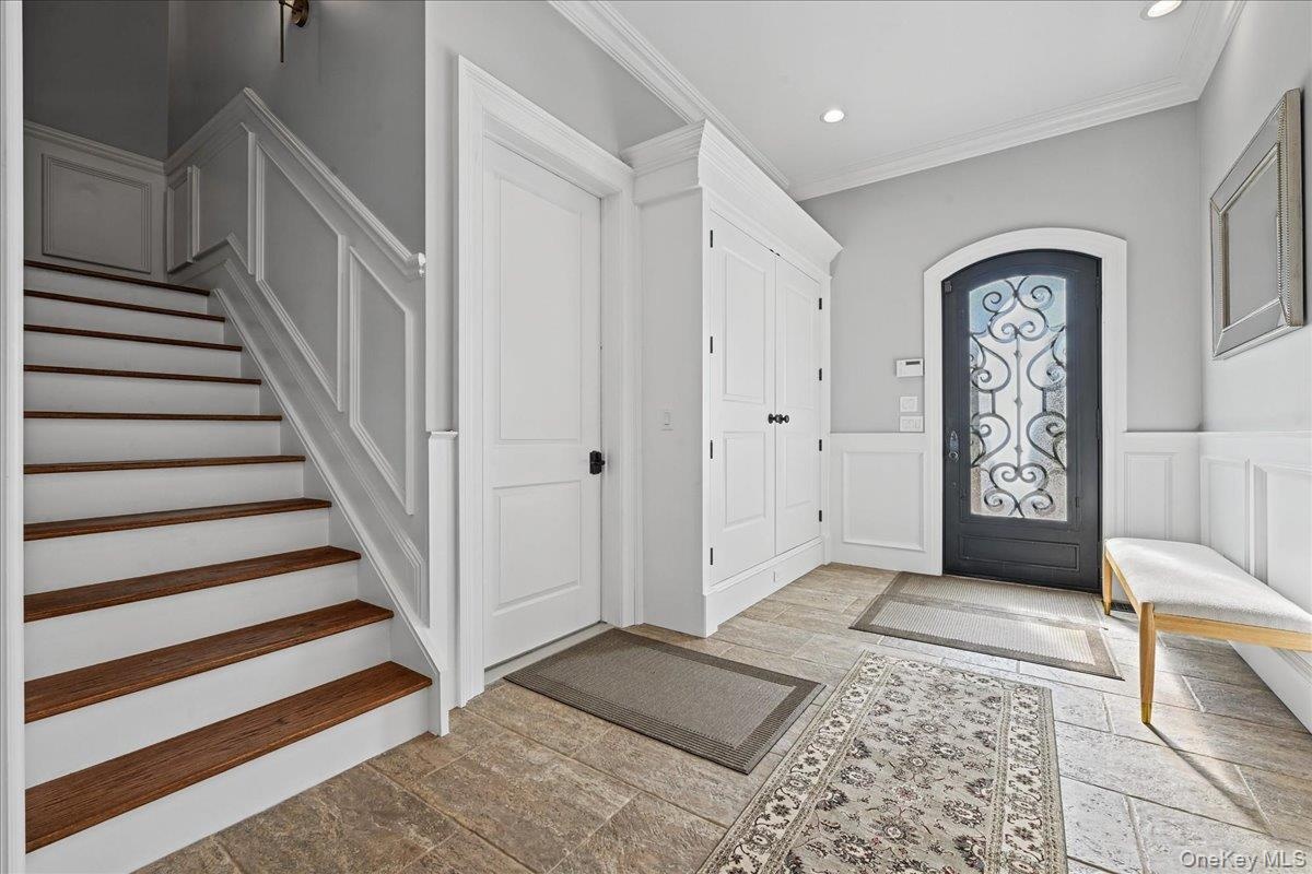 Entryway featuring tile flooring, wainscoting, an ornate arched entry door, a built-in storage cabinet, and a staircase with wood-finish treads