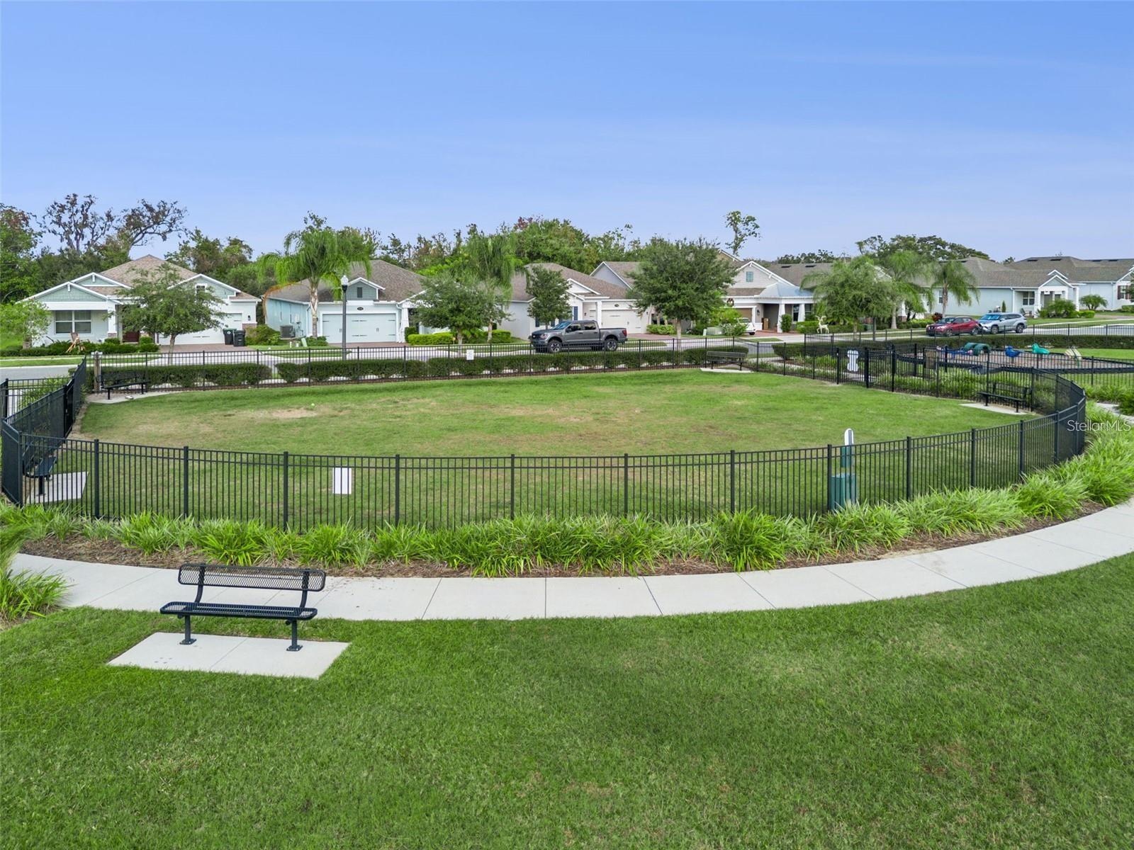 Fenced dog park with an open lawn, seating benches, and surrounding walking paths.