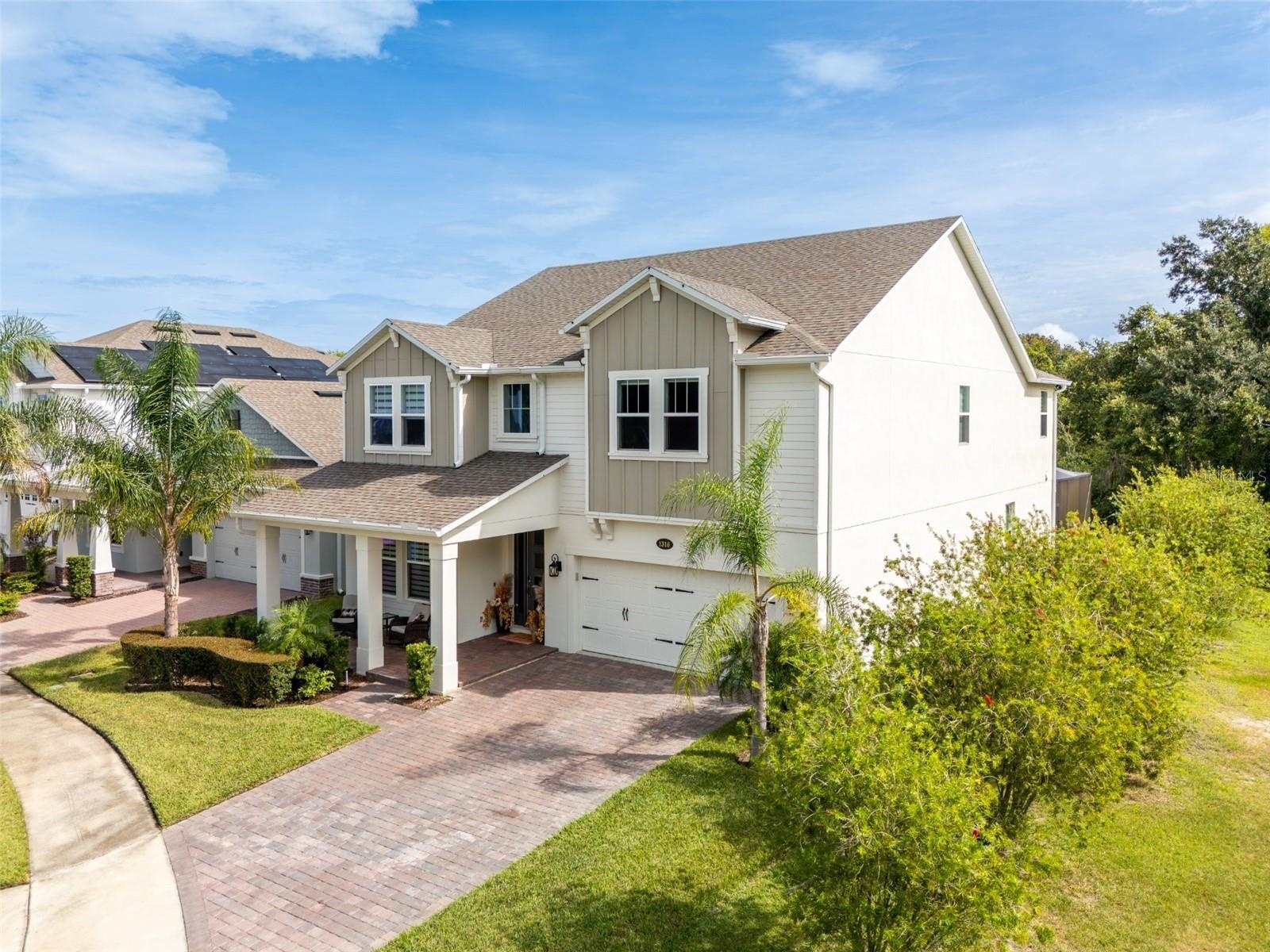 Elegant two-story home with a welcoming covered porch, paver driveway, and lush tropical landscaping.