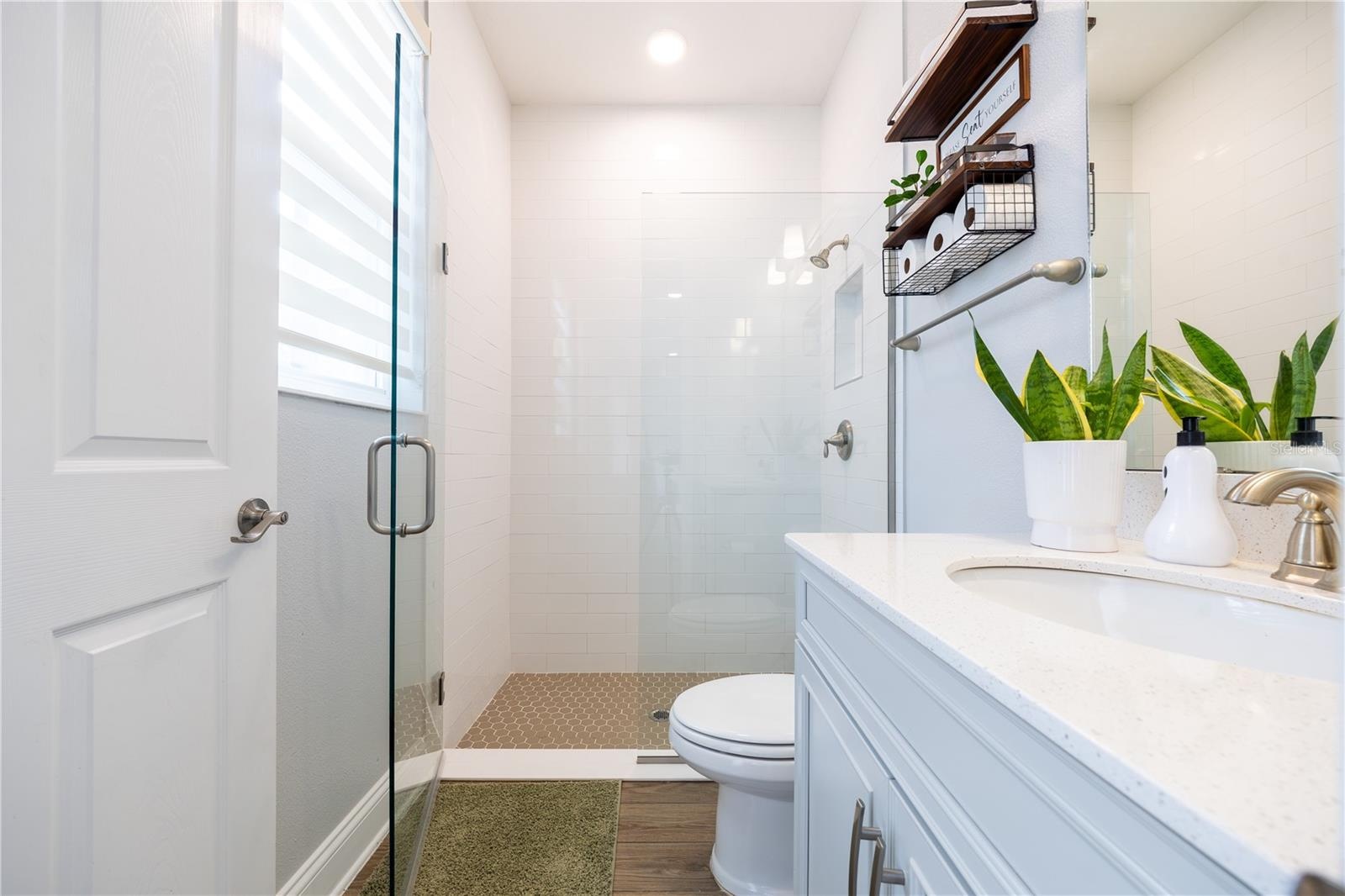 Bright bathroom featuring a frameless glass shower, white tile walls, mosaic shower floor, and a spacious vanity countertop.