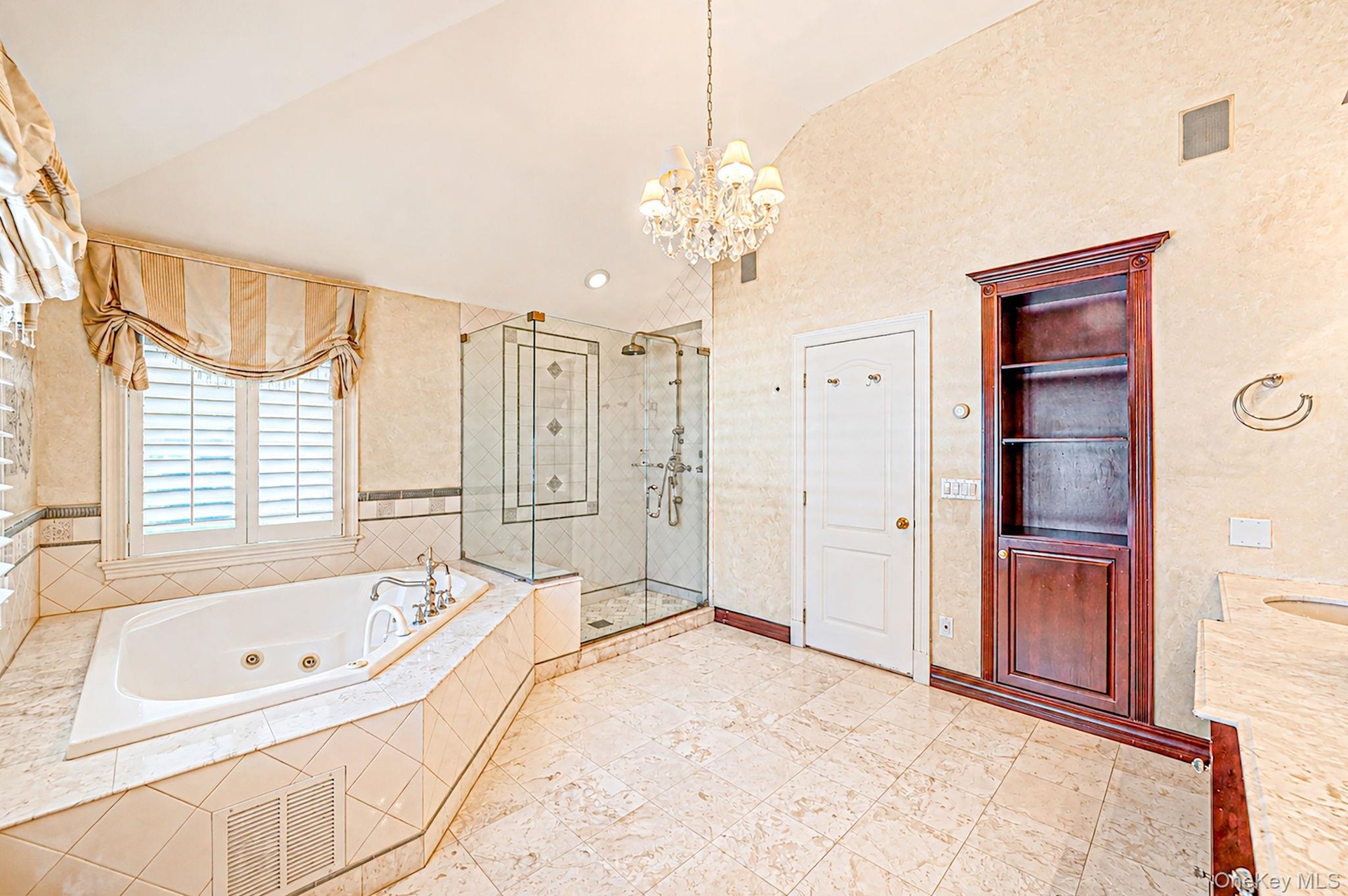 Jetted soaking tub with tile surround, glass-enclosed shower, built-in shelving, and chandelier lighting