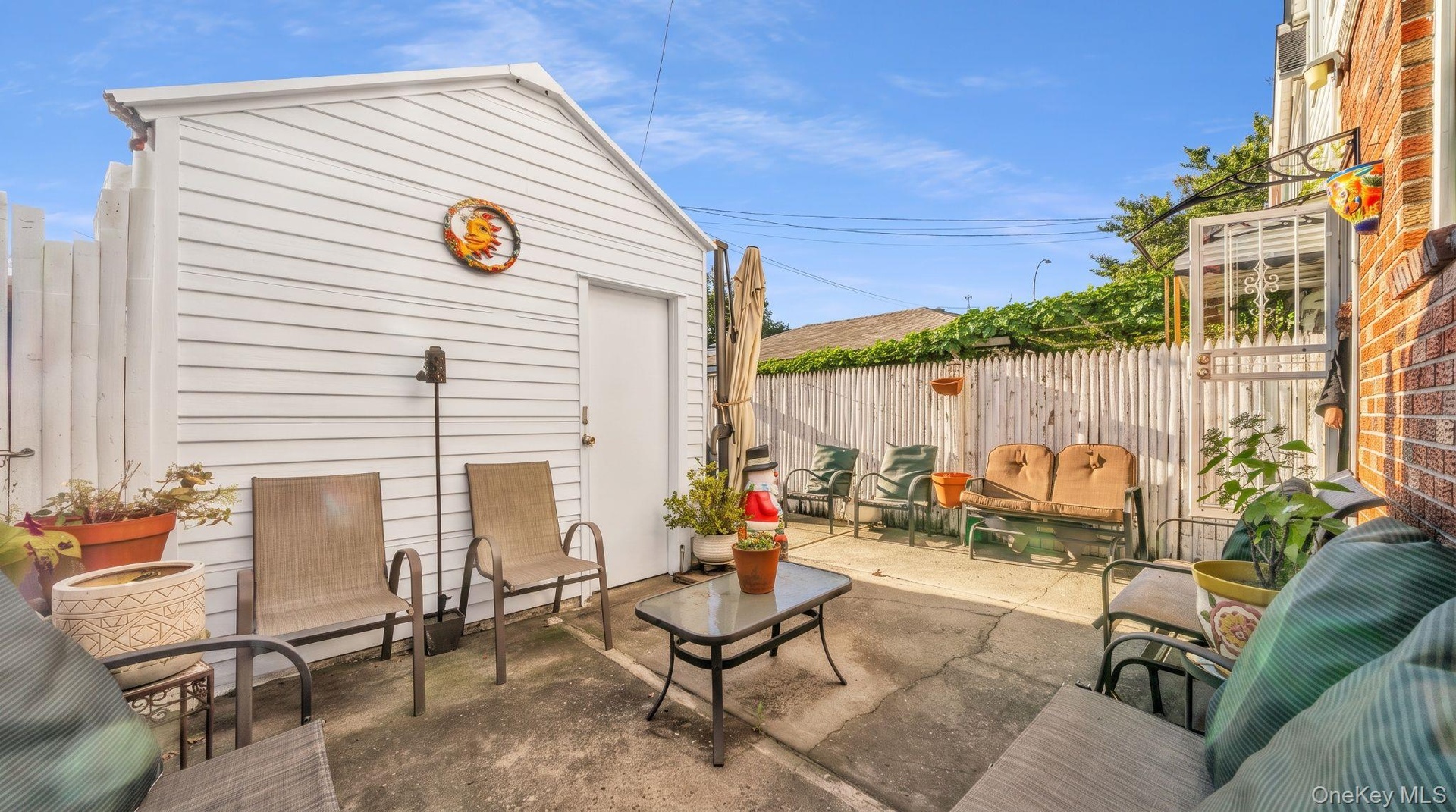 Paved patio area, white picket fencing, brick exterior wall, detached storage structure with siding, and a security door