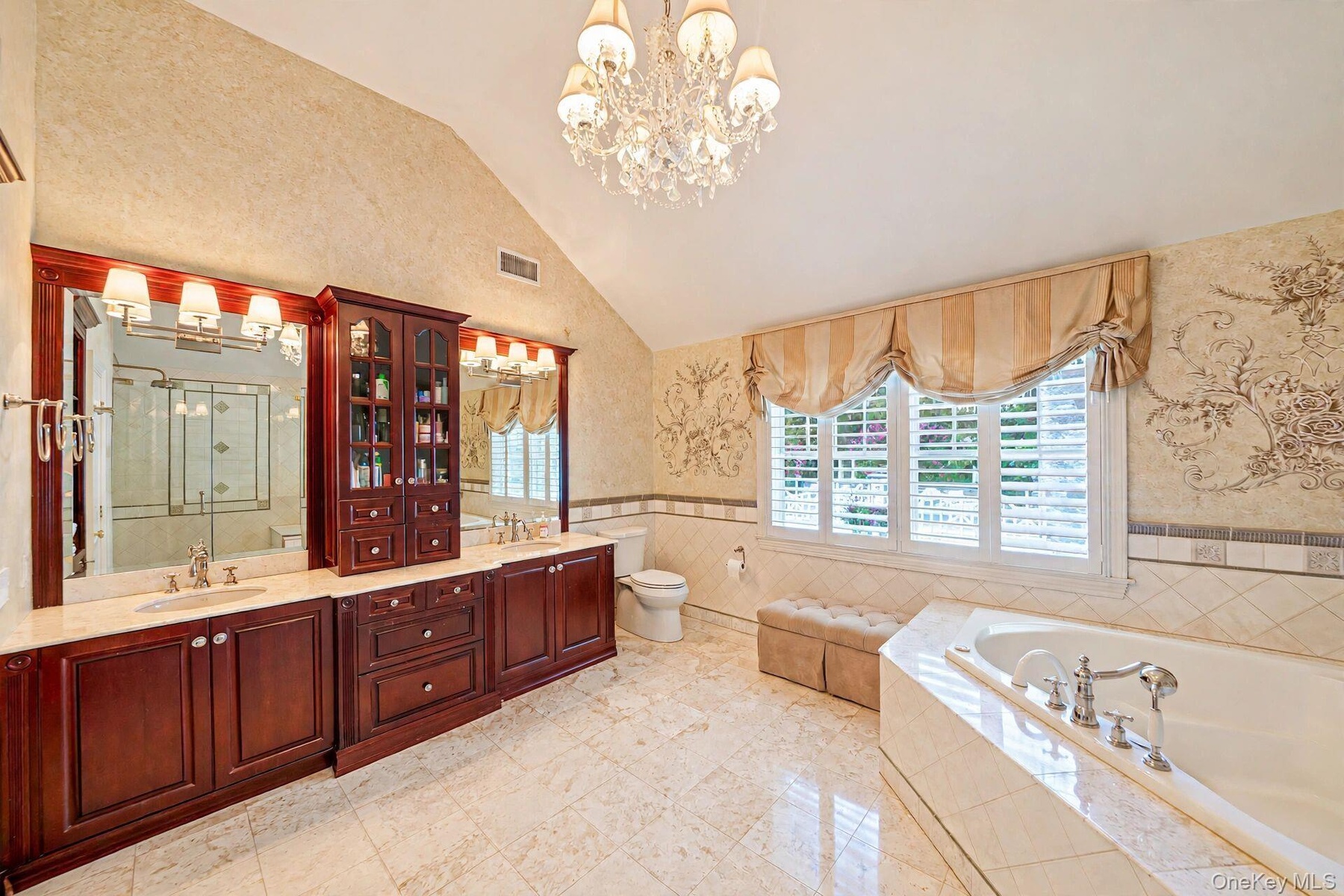 Vaulted ceiling with chandelier, extensive wood-finish cabinetry, dual vanity with undermount sinks, and a built-in tub