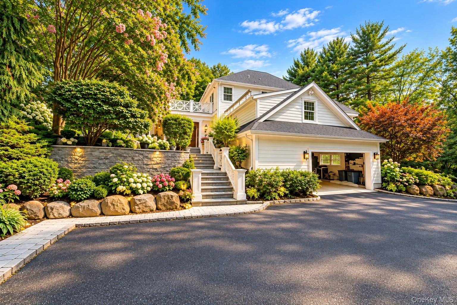 White siding exterior with a dark shingle roof, attached garage, stone retaining walls, stone steps, and mature landscaping