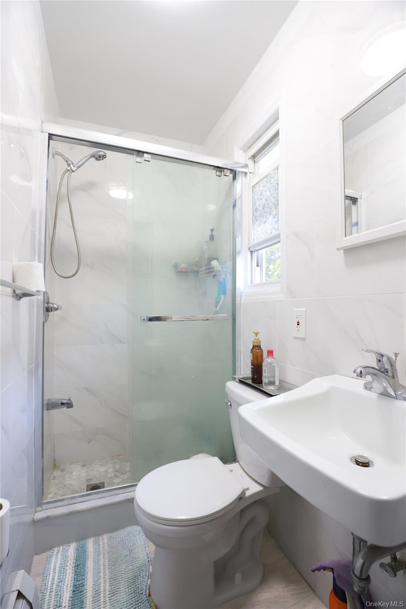White tiled bathroom featuring a glass-enclosed shower, wall-mounted sink, and a window