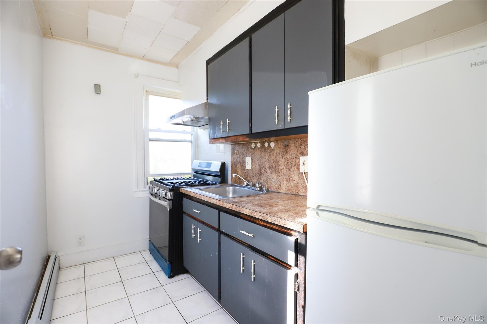 Galley kitchen featuring dark cabinetry with brushed metal hardware, wood-finish countertops, tile flooring, stainless steel range hood, and white app
