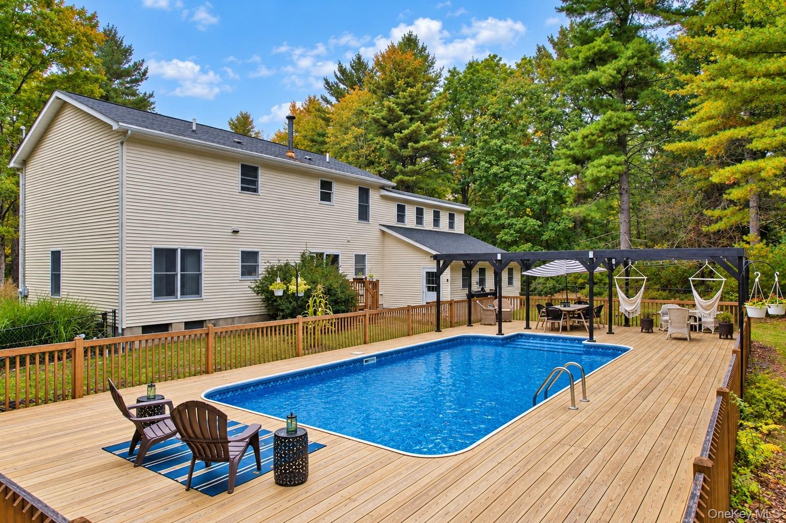Spacious wood-finish deck with an in-ground swimming pool, a black pergola, and a privacy fence, surrounded by mature trees