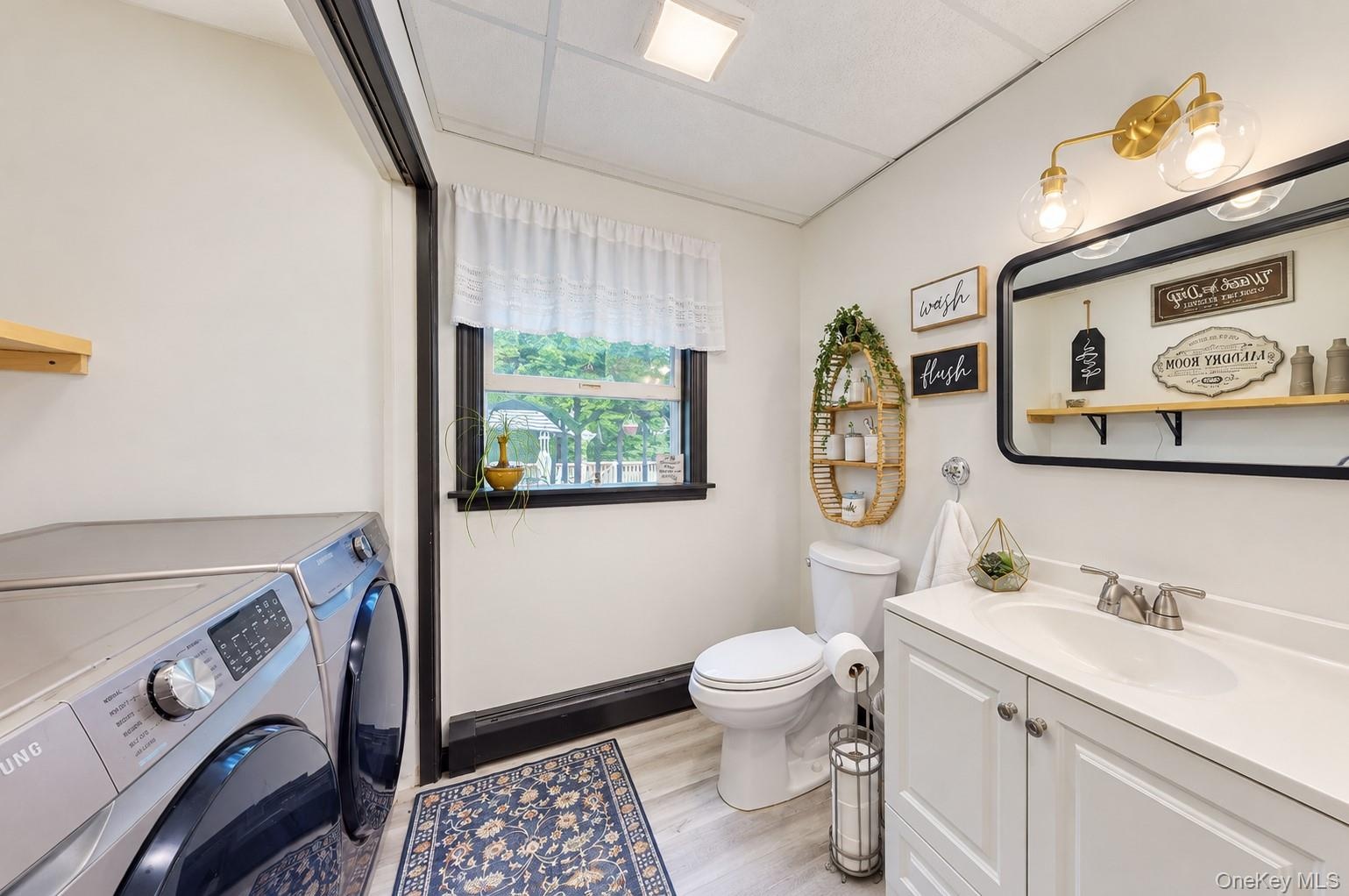 Light-toned wood-finish flooring, white vanity with integrated sink, rectangular framed mirror, and a brass-finish vanity light fixture