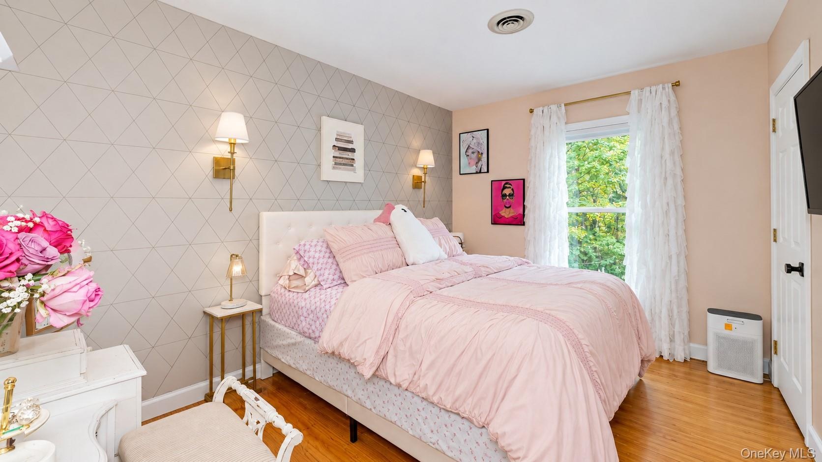 Hardwood flooring, geometric pattern accent wall, brass wall sconces, ceiling-mounted vent, and a single window with a gold-tone curtain rod
