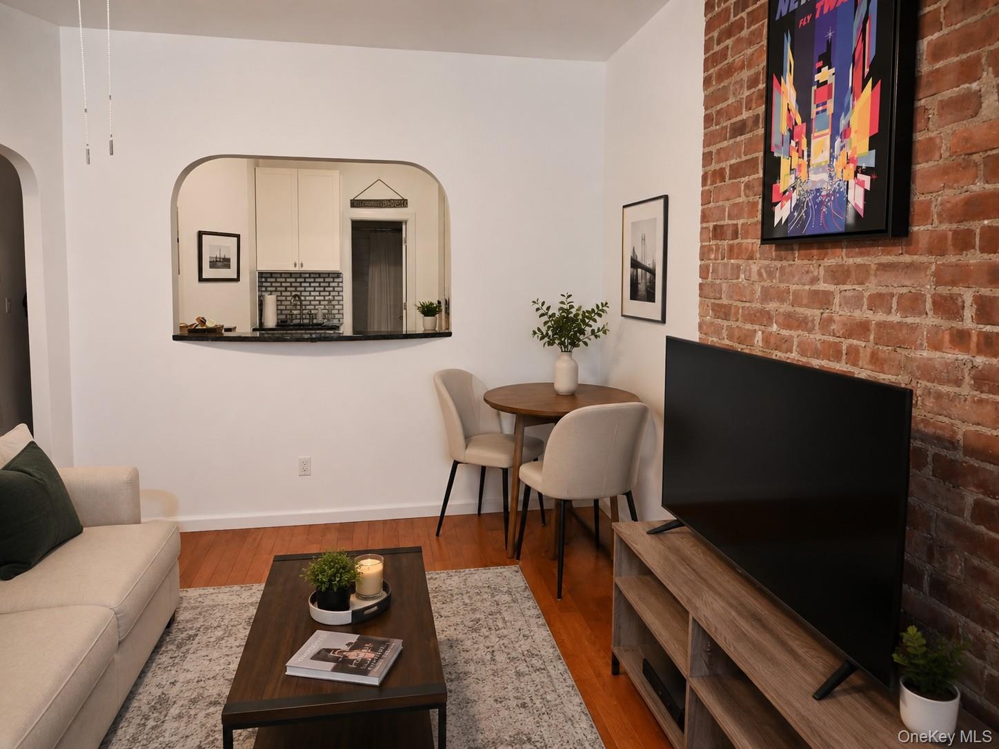 Exposed brick accent wall, wood-finish flooring, and a kitchen pass-through with a dark countertop (virtually staged)