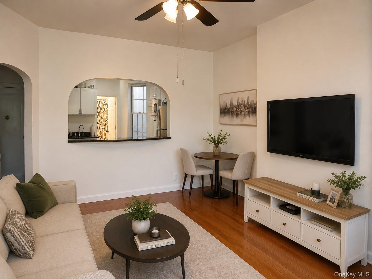 Open concept living area featuring wood-finish flooring, a ceiling fan with integrated lighting, and an arched pass-through window to the kitchen. (vi