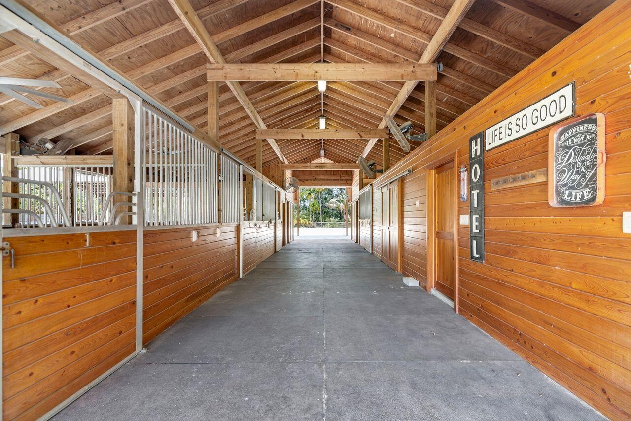 Spacious barn interior featuring exposed wood beams and ceiling, multiple stalls with custom stall doors, center isle, fans, fly system, 3 stalls have