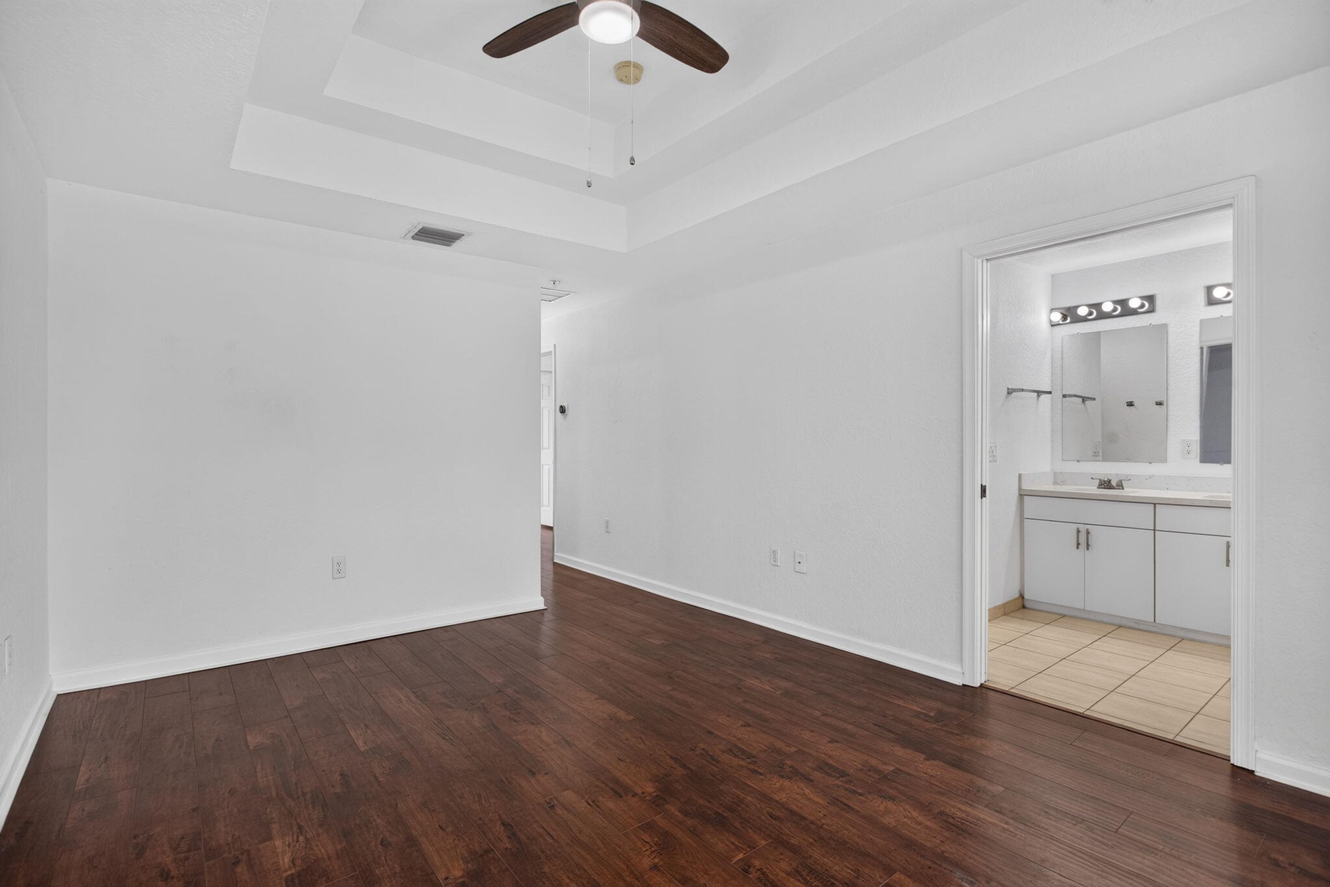 Wood-finish flooring, tray ceiling, ceiling fan, and an en-suite bathroom featuring a double vanity with an integrated mirror