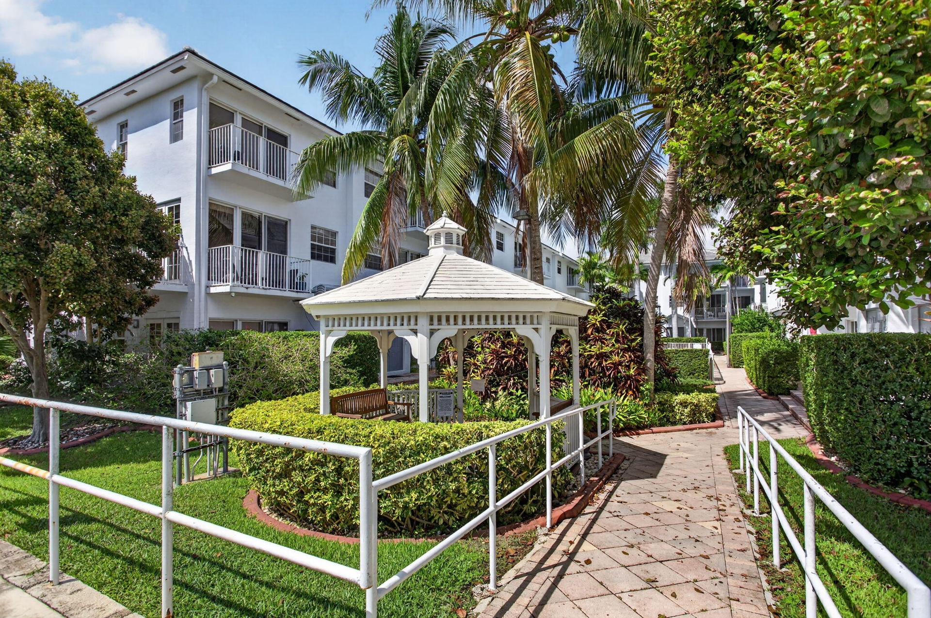 Multi-level building with individual balconies, white gazebo, paved pathway, manicured hedges, and mature palm trees