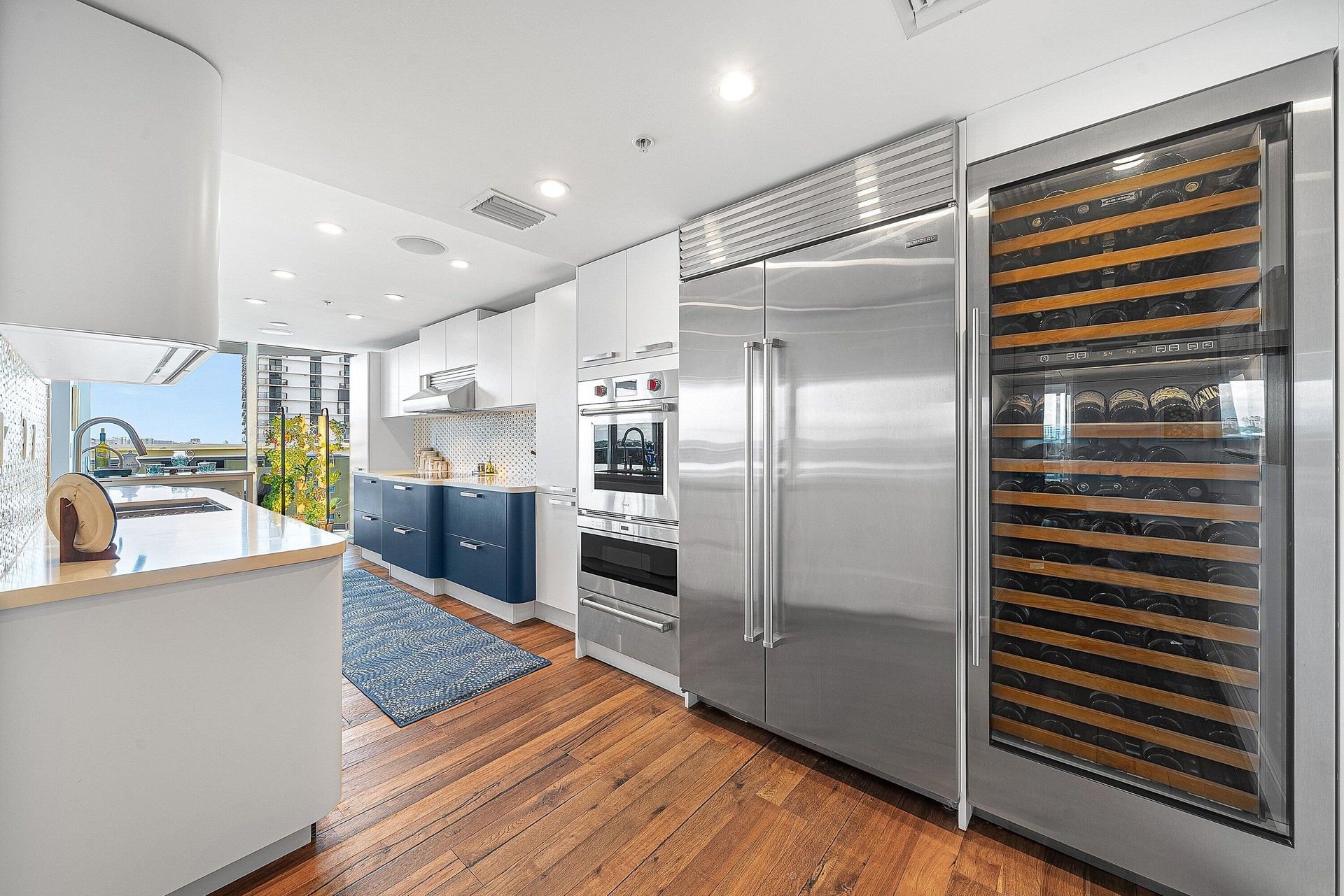 Gourmet kitchen featuring wood-finish flooring, stainless steel appliances, built-in wine refrigeration, white upper cabinetry, and contrasting blue l