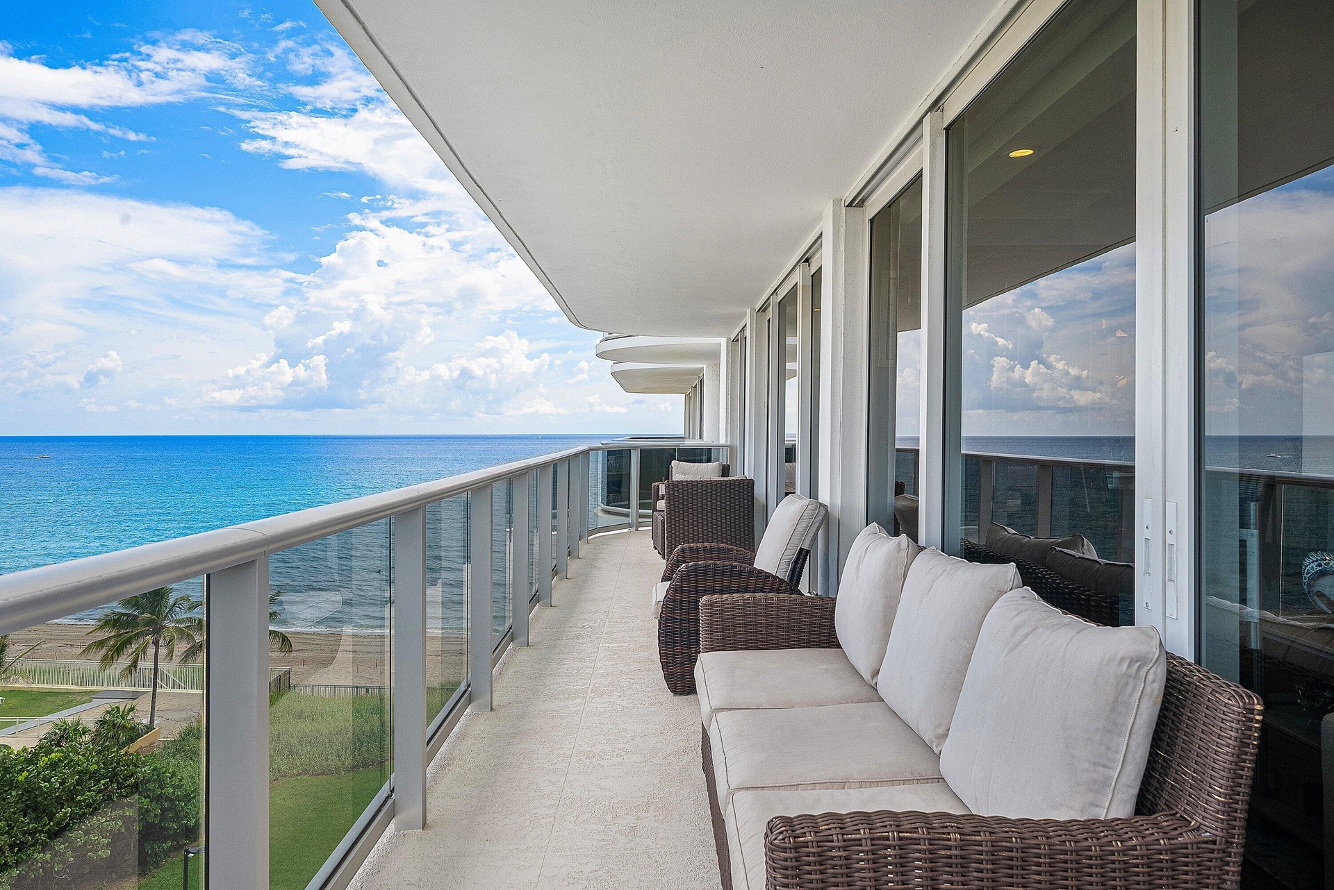 Expansive outdoor balcony with glass and metal railing, offering ocean scenery, a sand beach, and a grassy area