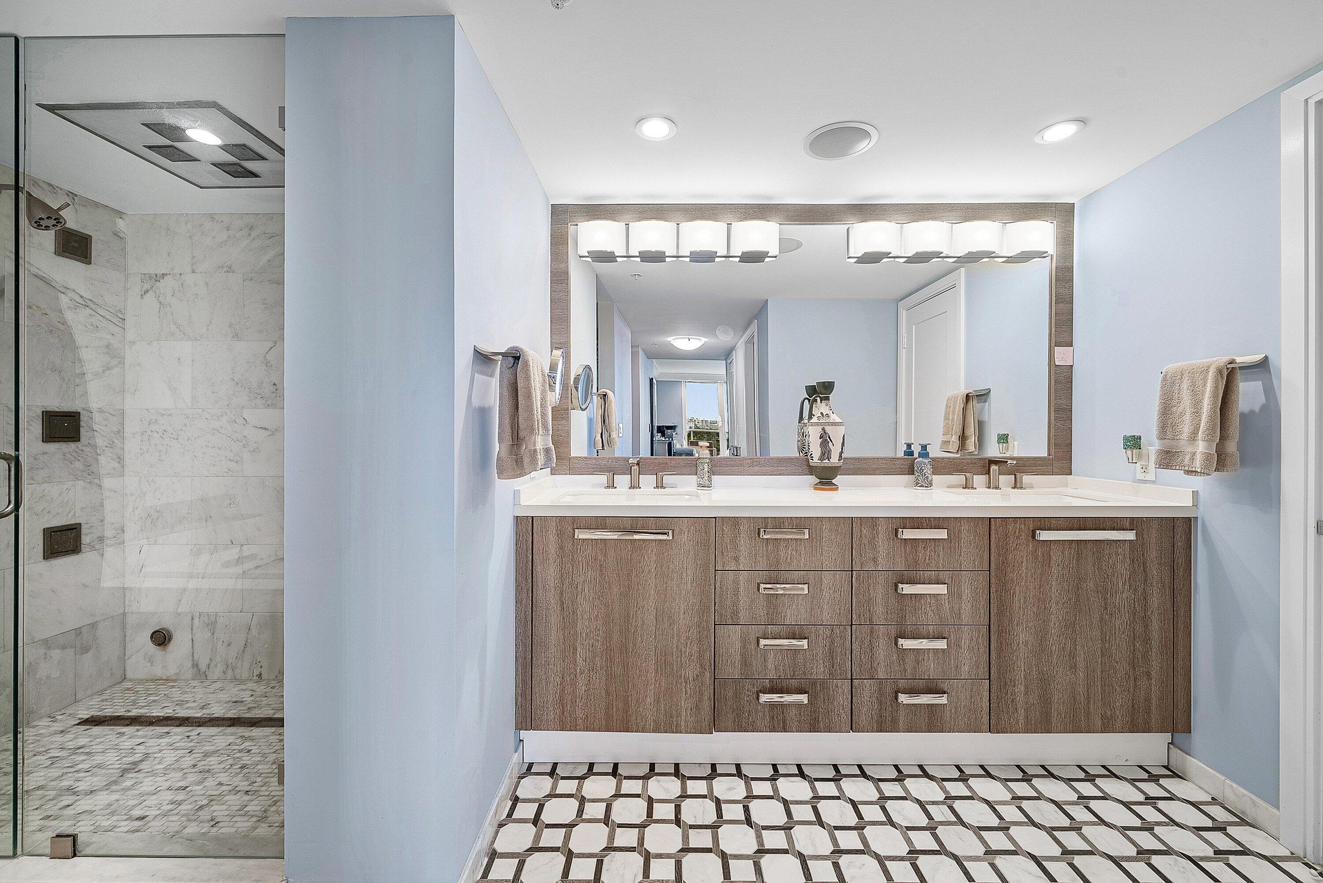 Glass-enclosed shower with marble-style tile, dual vanity with wood-finish cabinetry, stone countertop, and patterned floor tile