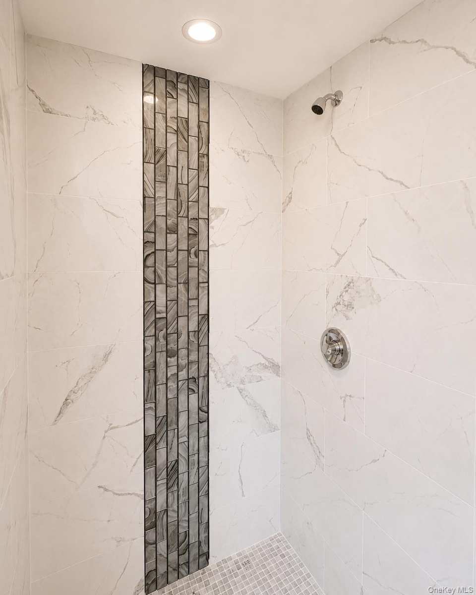 Shower enclosure featuring large format white tile walls with gray veining, a recessed ceiling light, and a vertical mosaic accent strip