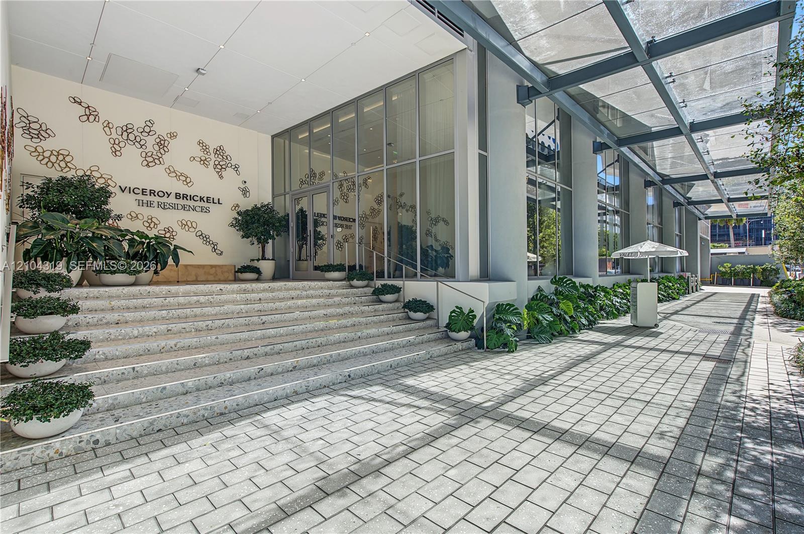 Building exterior featuring a glass curtain wall, expansive paver walkway, overhead steel-frame canopy, and integrated wide-tread stairs with decorati