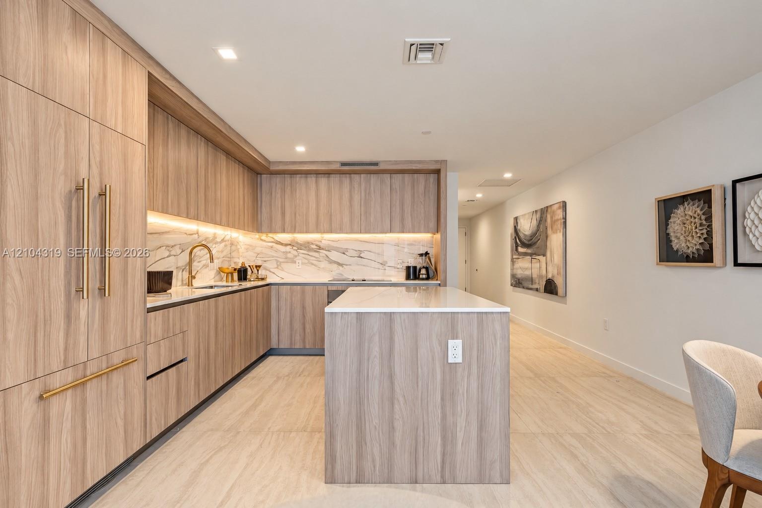 Kitchen featuring wood-finish cabinetry, a waterfall-edge island, stone-patterned backsplash, recessed lighting, and metallic hardware