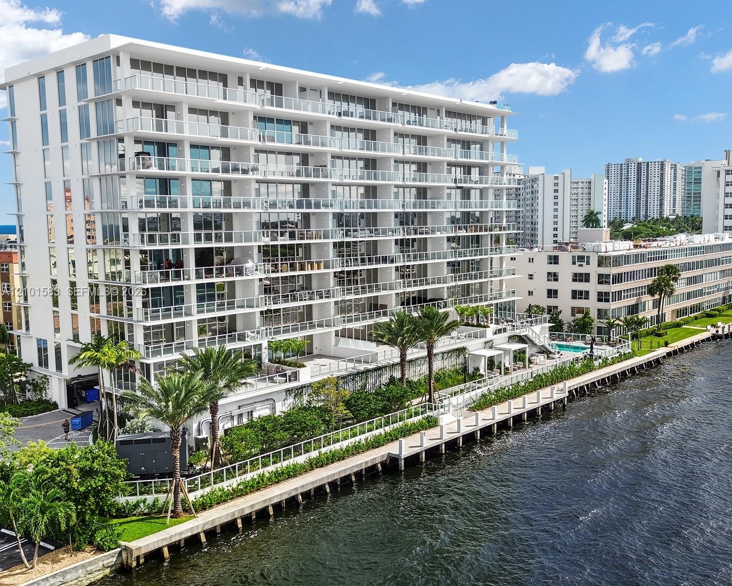 Modern mid-rise building featuring floor-to-ceiling glass windows, wraparound glass-panel balconies, a waterfront bulkhead, and a paved canal-side wal