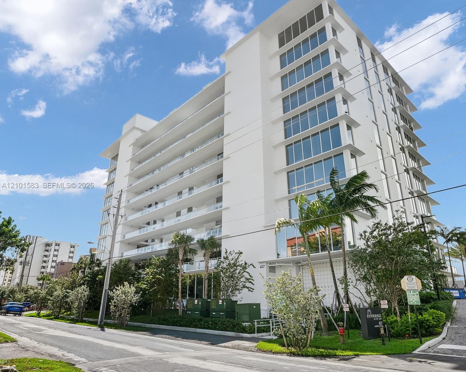 Modern mid-rise building exterior featuring a white stucco facade, expansive glass windows, and multiple stories of private balconies with glass raili
