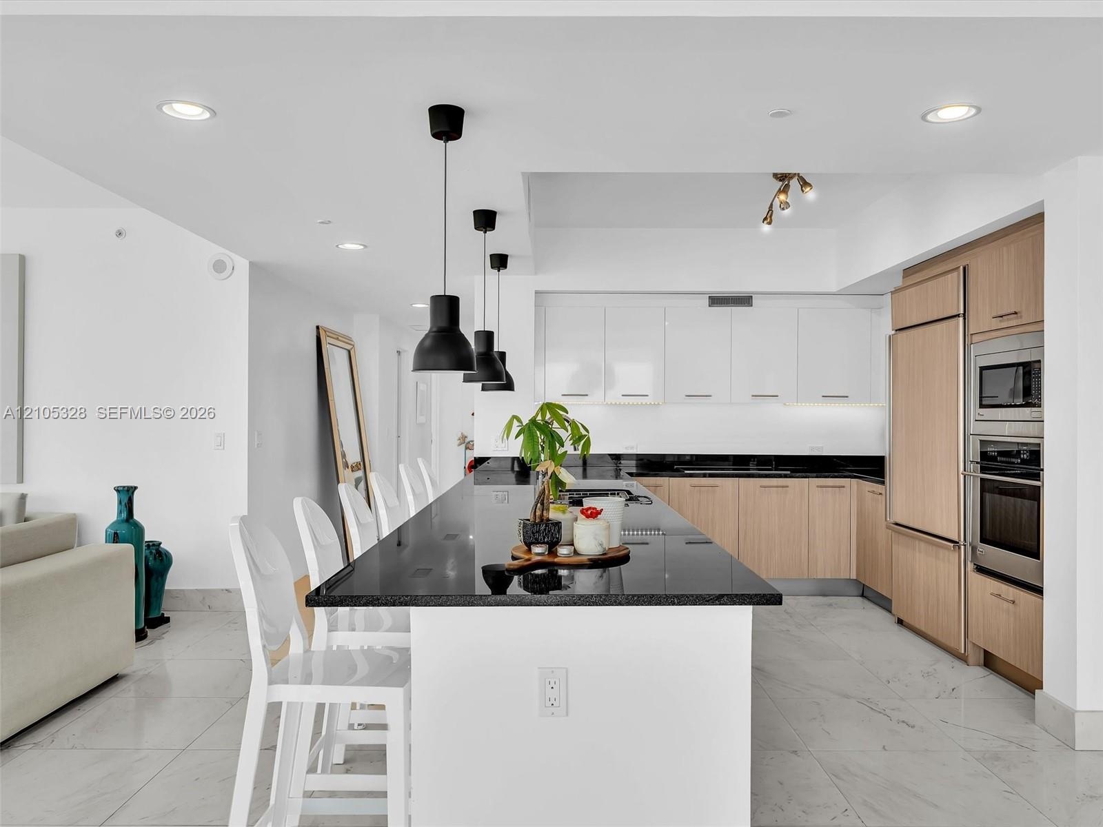 Modern gourmet kitchen featuring an oversized center island with seating, sleek dark stone countertops, stainless steel appliances, and wood-finish ca