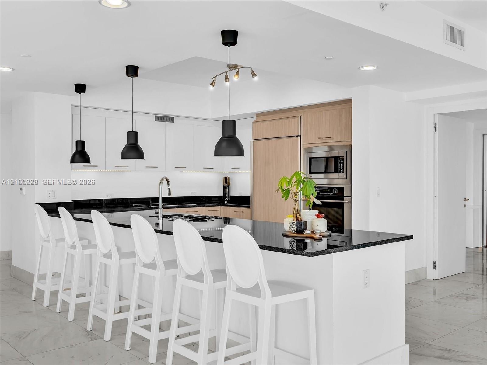 Modern gourmet kitchen featuring an oversized center island with seating, sleek dark stone countertops, stainless steel appliances, and wood-finish ca