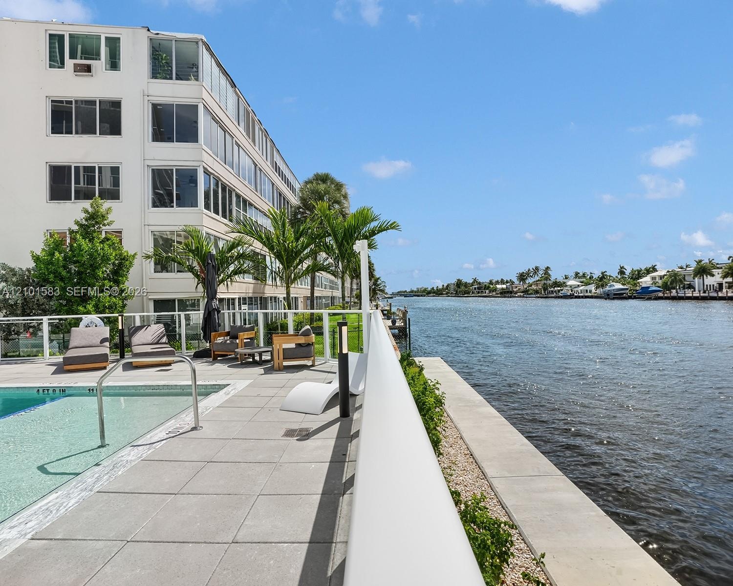 Waterfront pool area featuring expansive stone tile decking, a glass panel railing system, and manicured landscaping