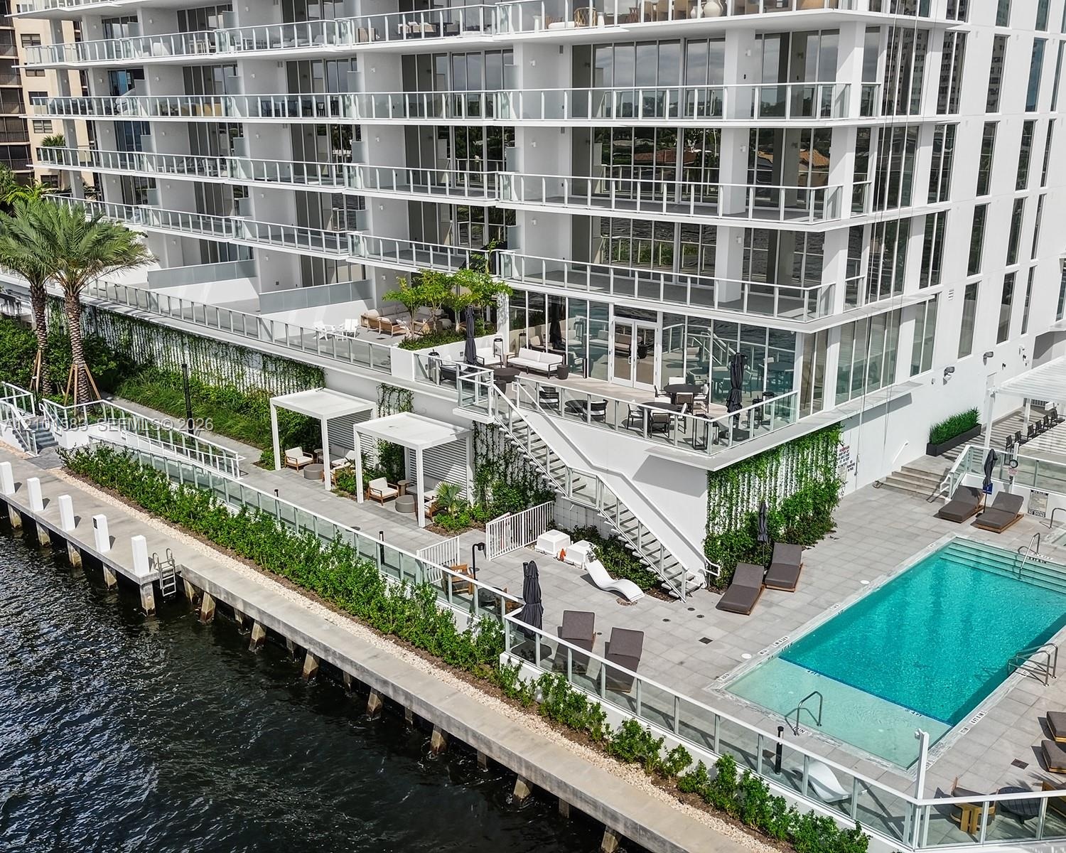High-rise building exterior featuring tiered balconies with glass railings, a waterfront pool deck, stone-paved sun terrace, integrated pergolas, and 